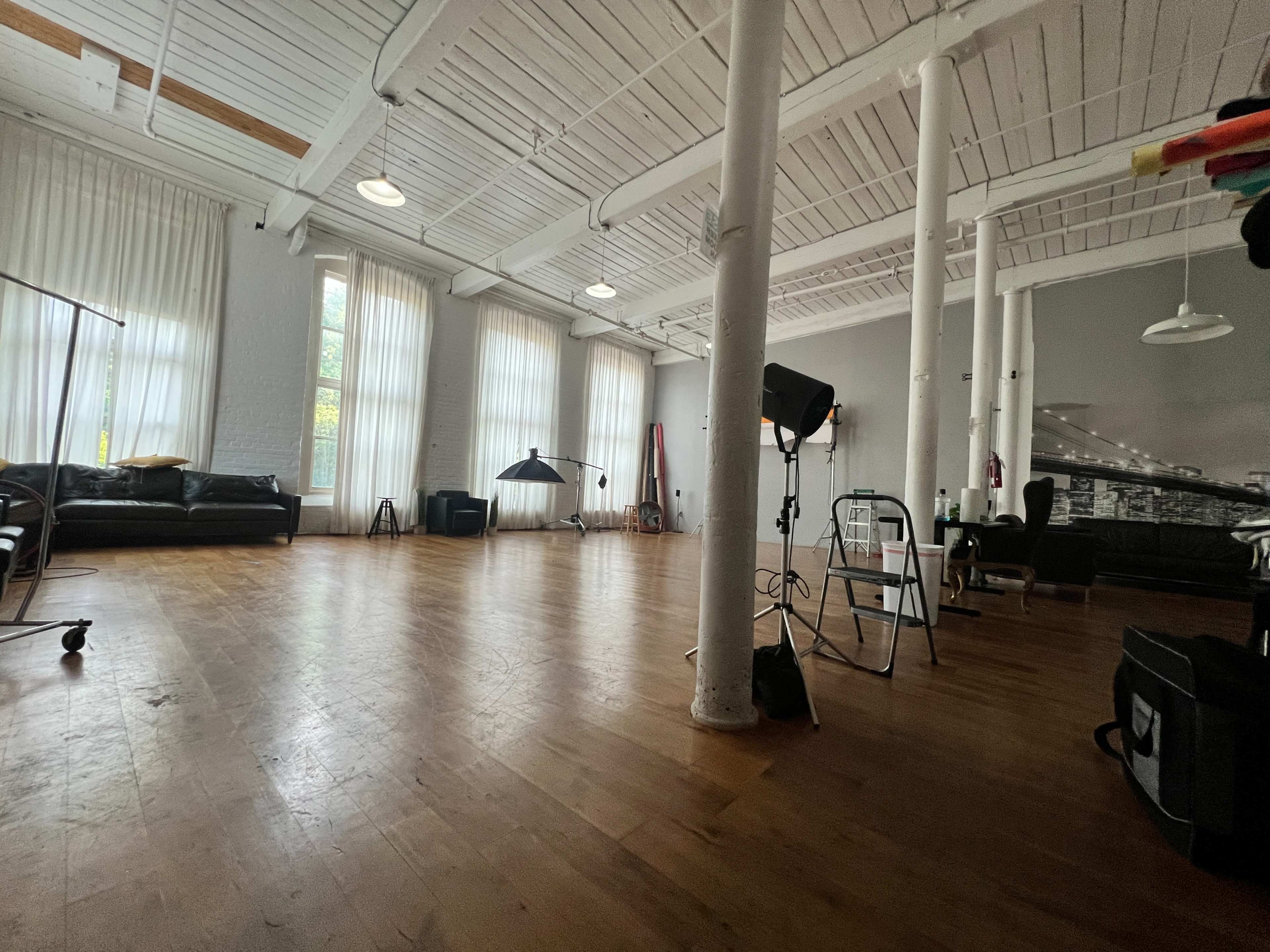The image shows a spacious, well-lit studio with wooden flooring, large windows, and minimal furniture, including a couple of black sofas and lighting equipment.