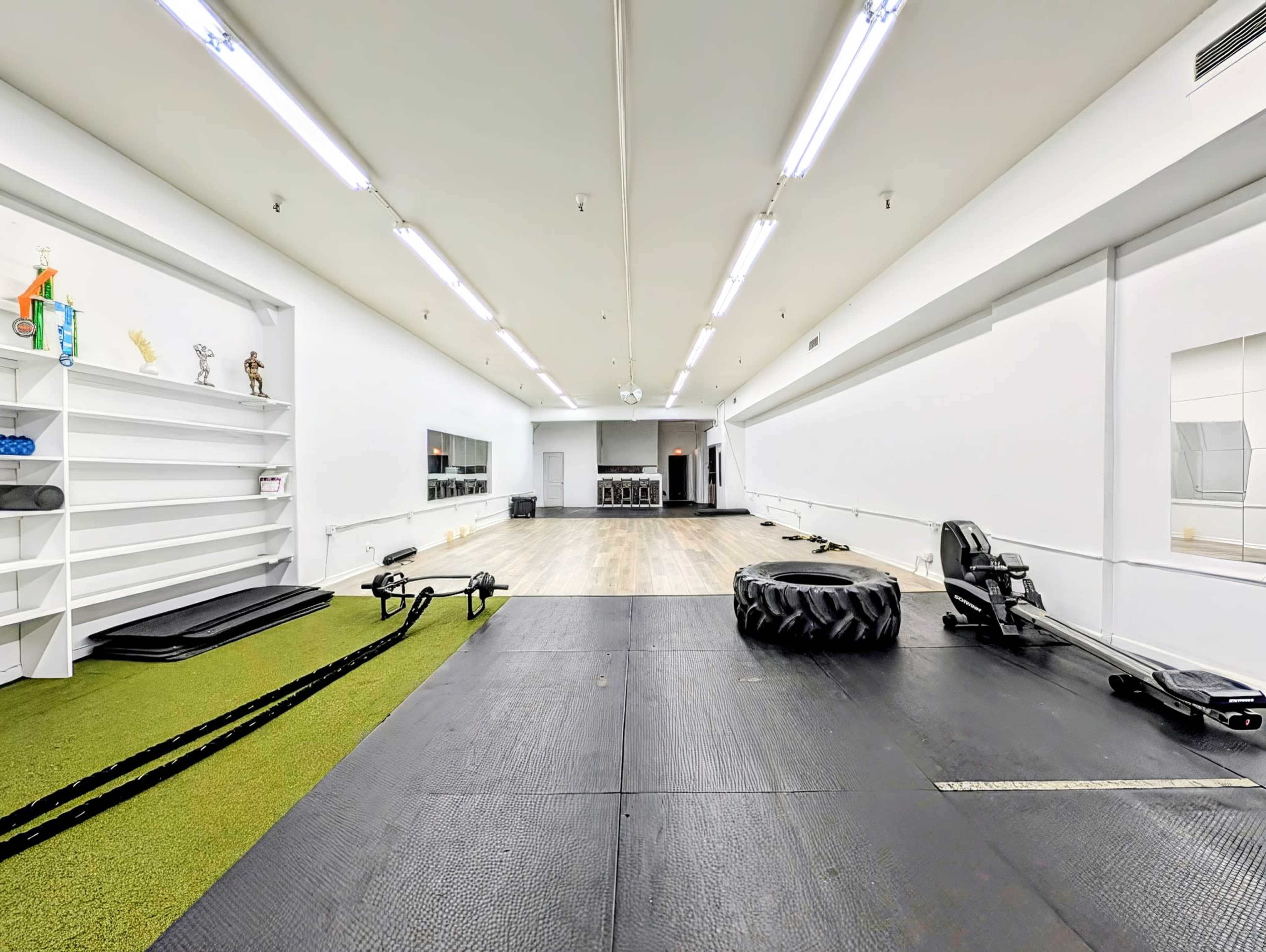 The image shows a spacious gym area featuring a black rubber floor, exercise equipment, and a mirrored wall, with a green turf section on one side.