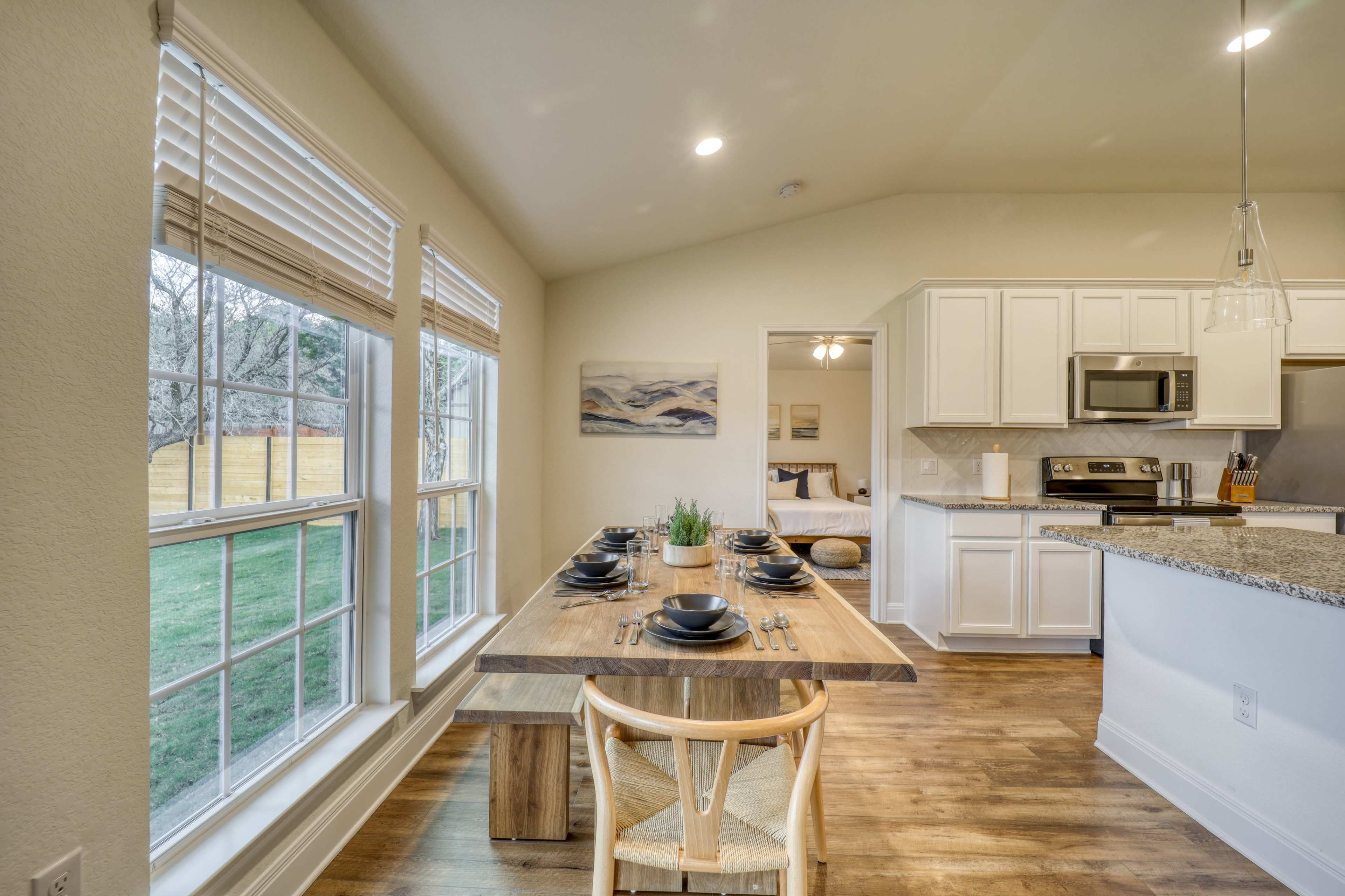 The image shows a bright dining area featuring a wooden table set for eight, with large windows overlooking a grass lawn and a modern kitchen in the background.