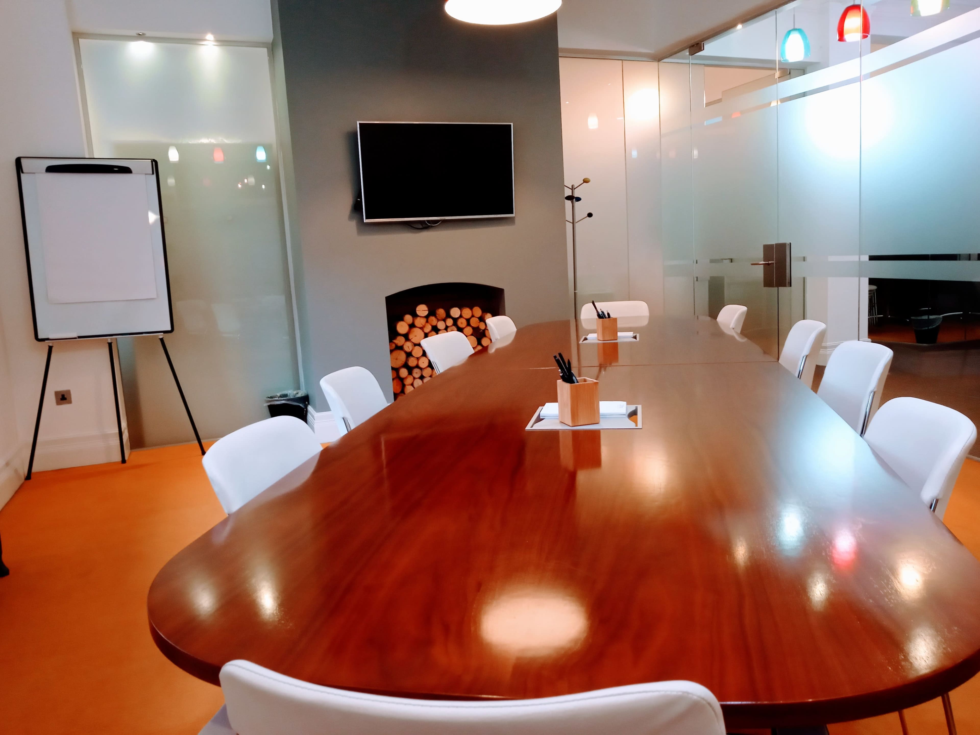 A modern conference room featuring a long wooden table, white chairs, a television on the wall, and a stack of logs in a decorative fireplace.