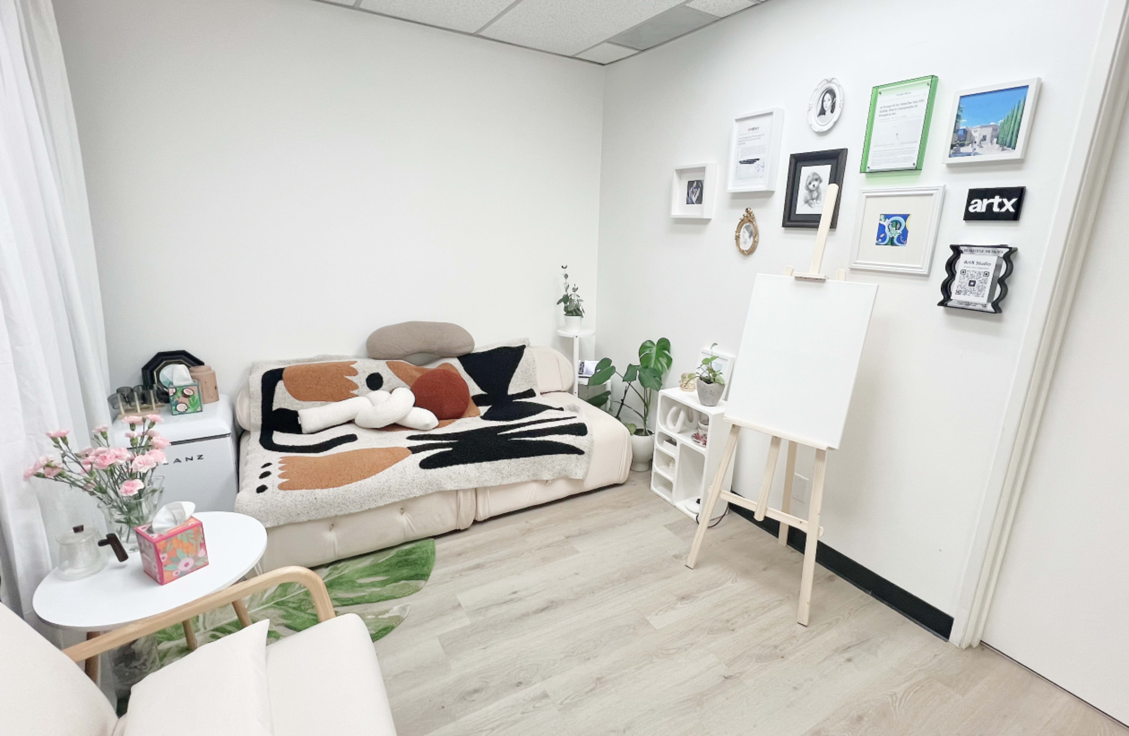The image shows a cozy room featuring a sofa with a patterned throw, an easel, and a wall adorned with framed pictures and certificates.