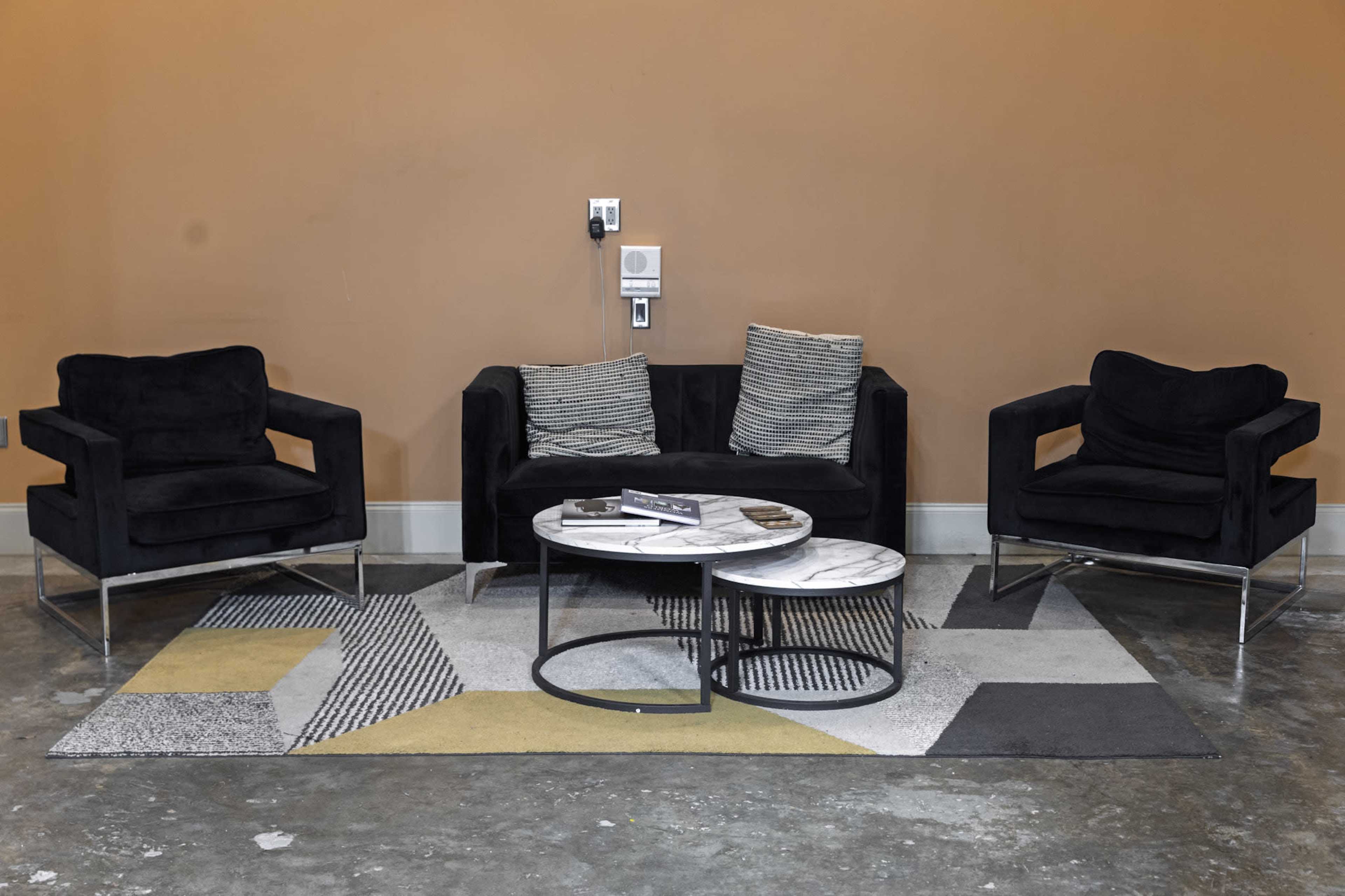The image shows a modern living room setup featuring a black upholstered couch and two matching armchairs, along with a round coffee table and a geometric patterned area rug.