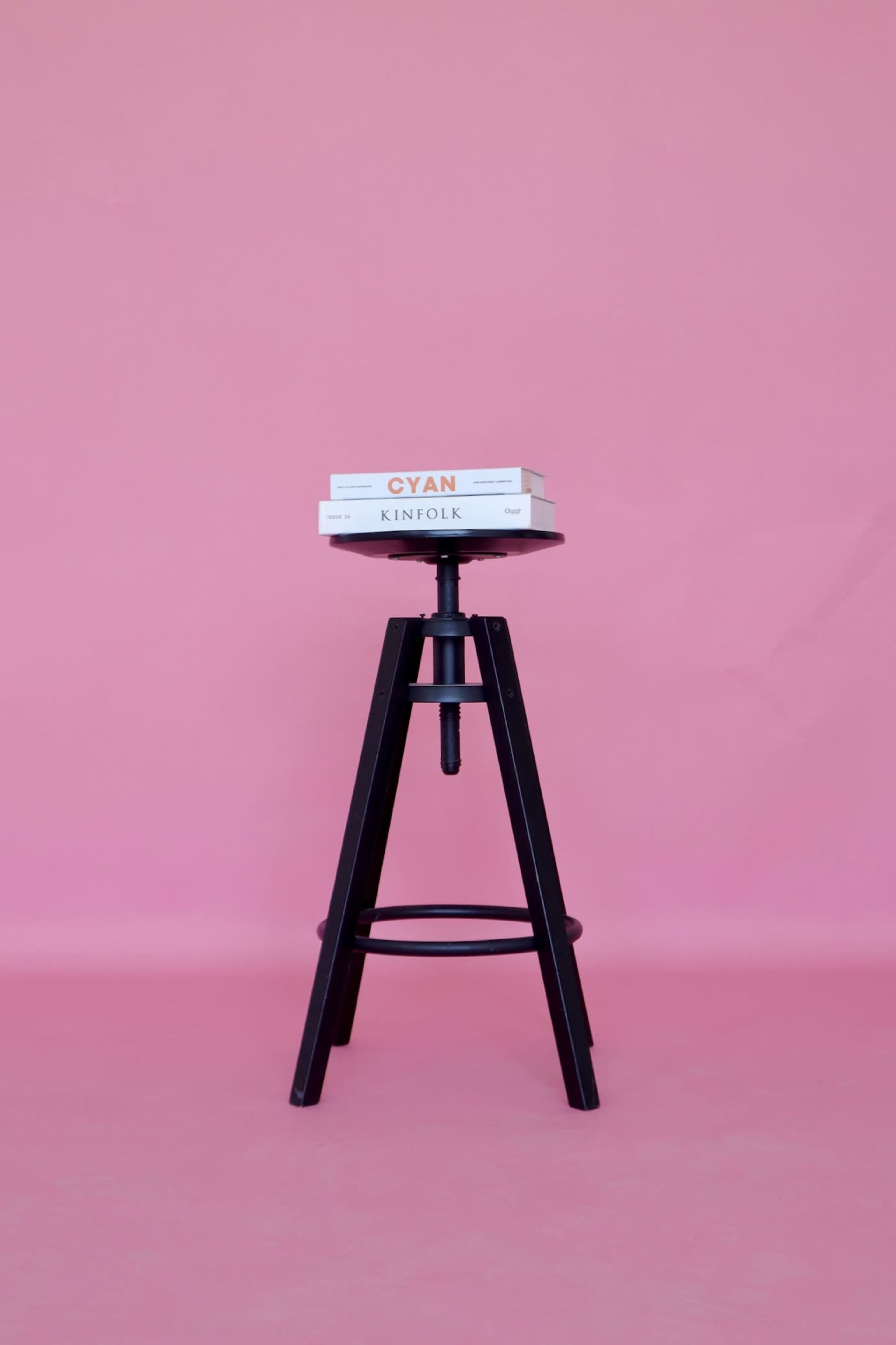 A black stool with a height-adjustable seat holds two stacked books against a pink background.