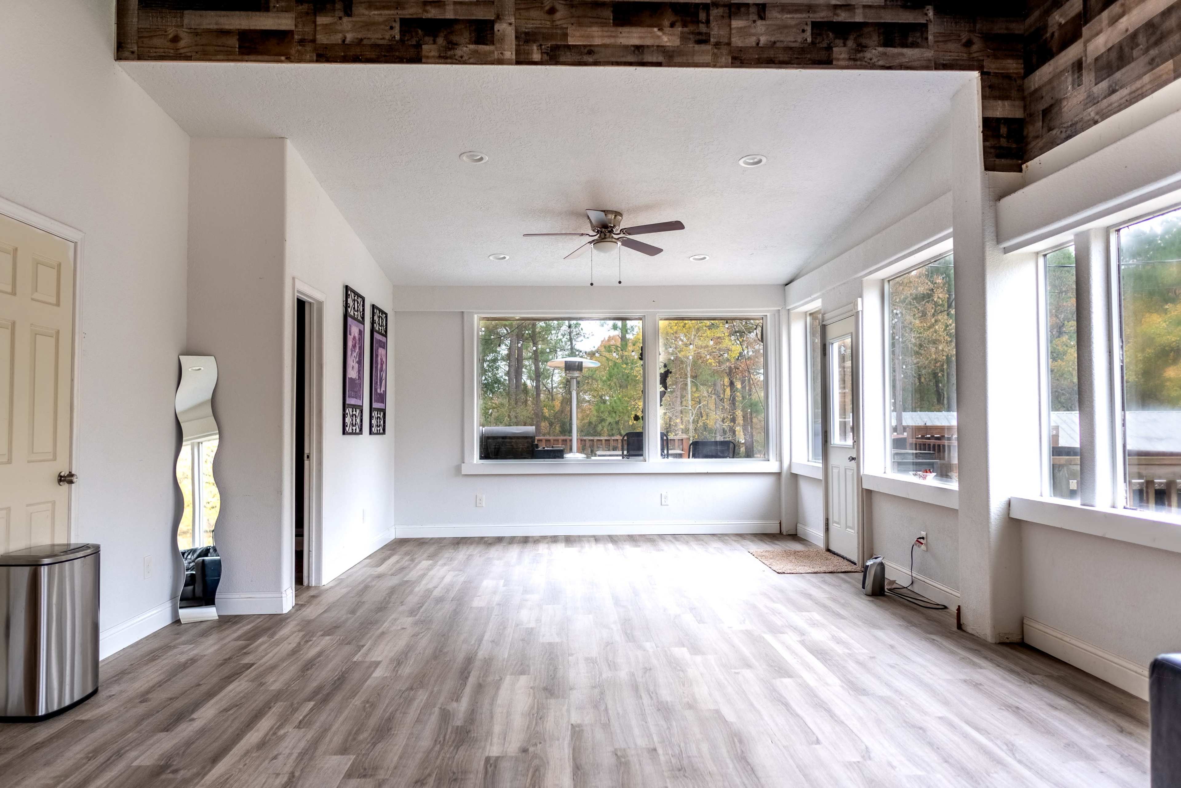 The image shows a spacious, bright interior room with large windows, a ceiling fan, and light-colored wooden flooring.