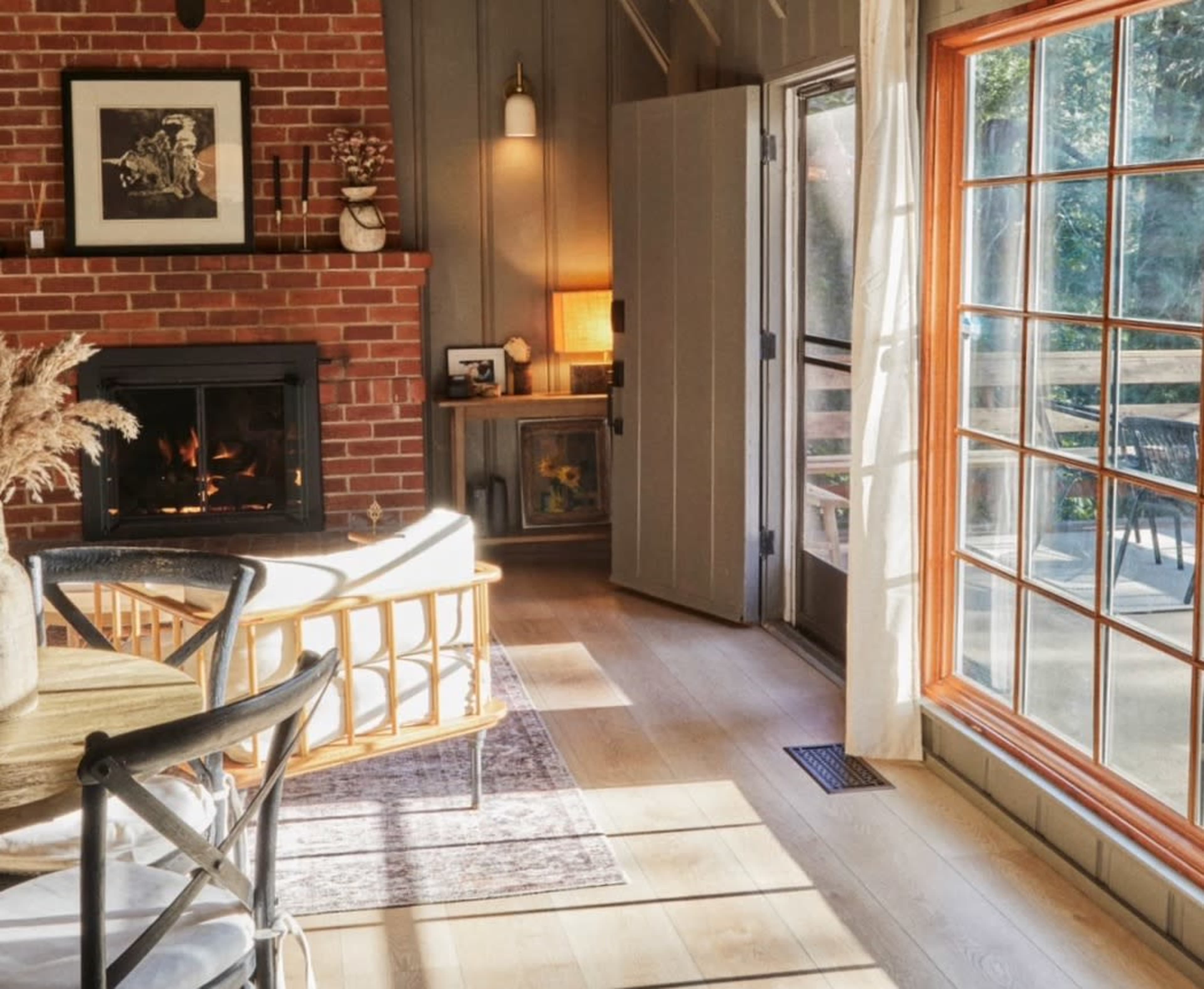 A cozy living area with a brick fireplace, a rattan chair, and large windows allowing natural light to flood the space.