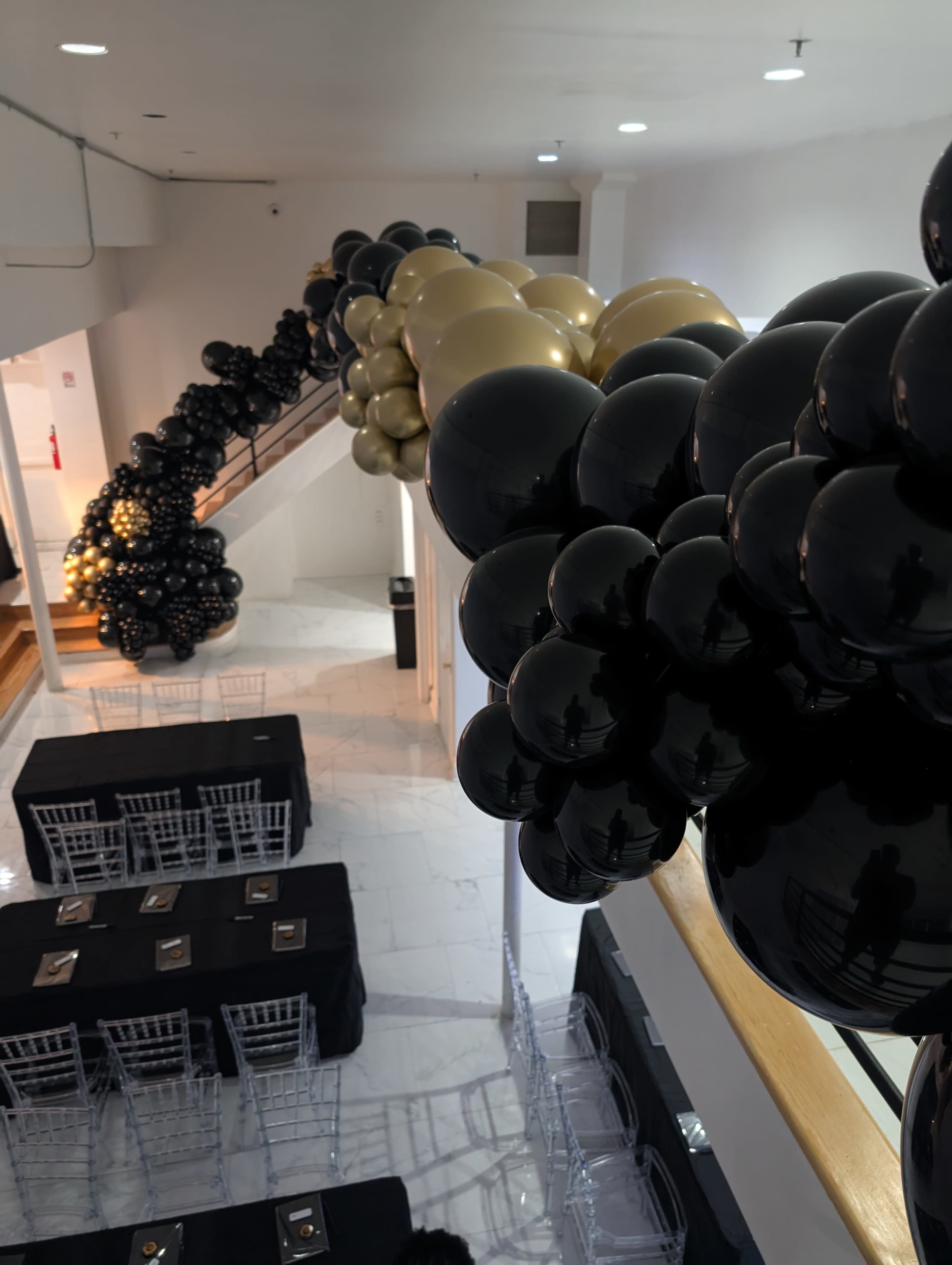 A staircase adorned with a decorative balloon garland in black, gold, and clear colors, above tables set for an event in a modern venue.