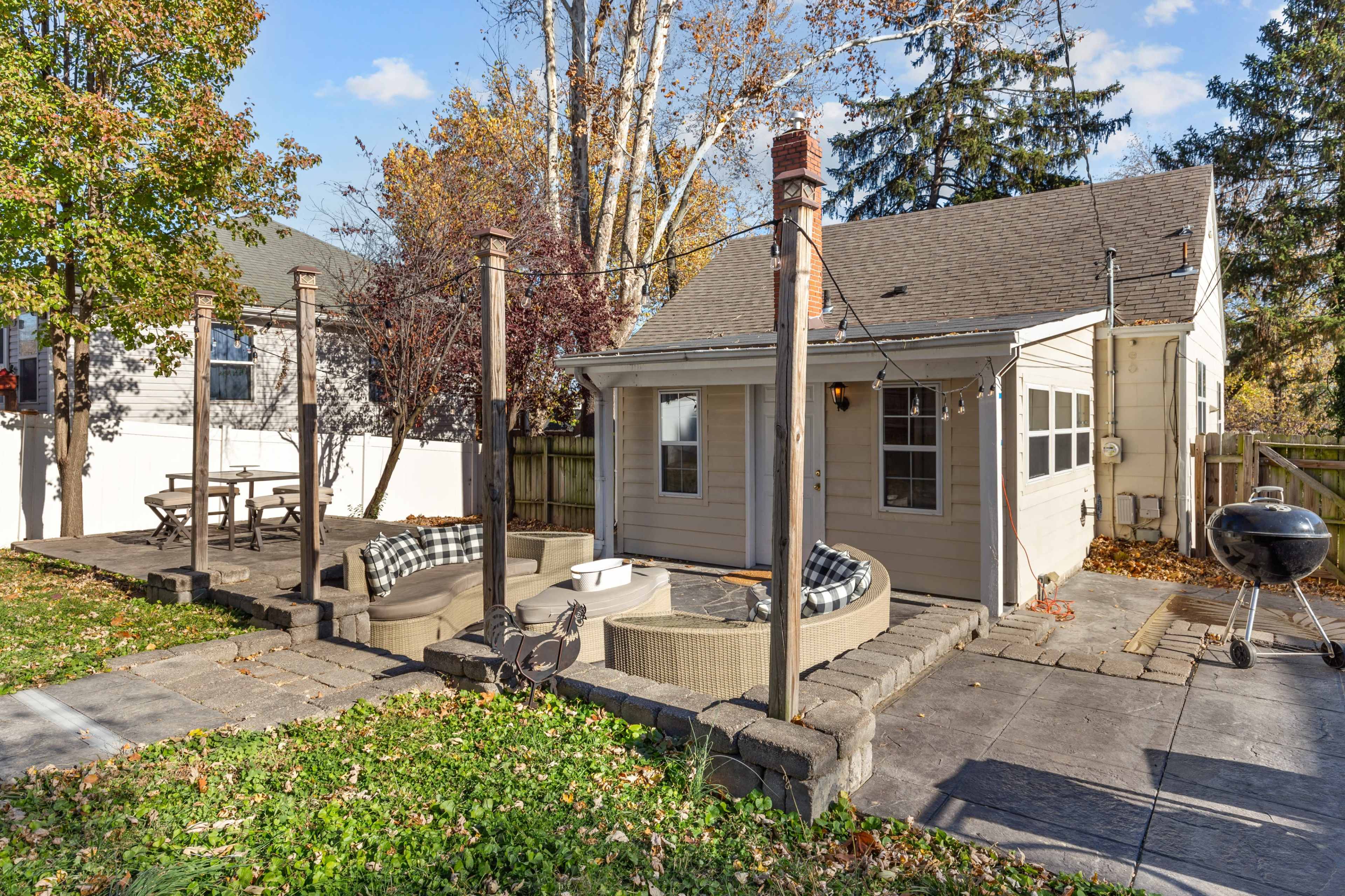 The image shows a backyard with a patio area featuring a fire pit and seating, surrounded by trees and a house in the background.