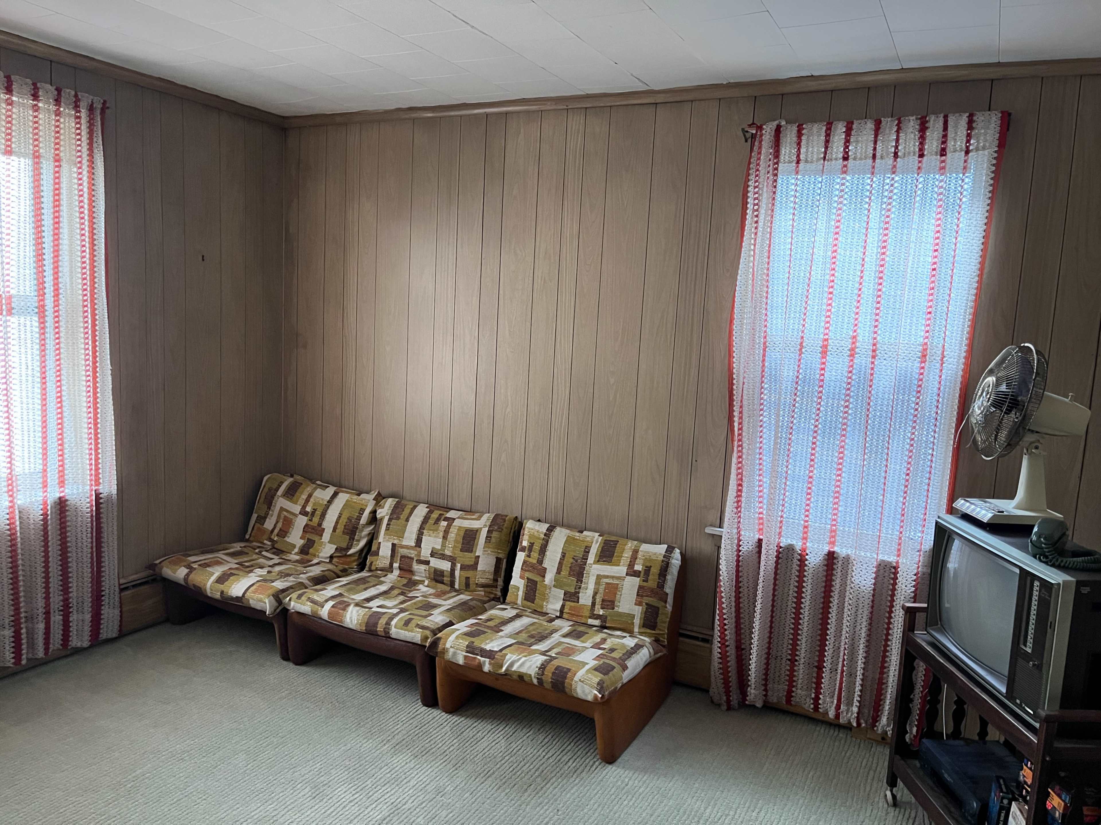 The image shows a small living room with a patterned sofa, two windows covered in striped curtains, and a television set on a stand.