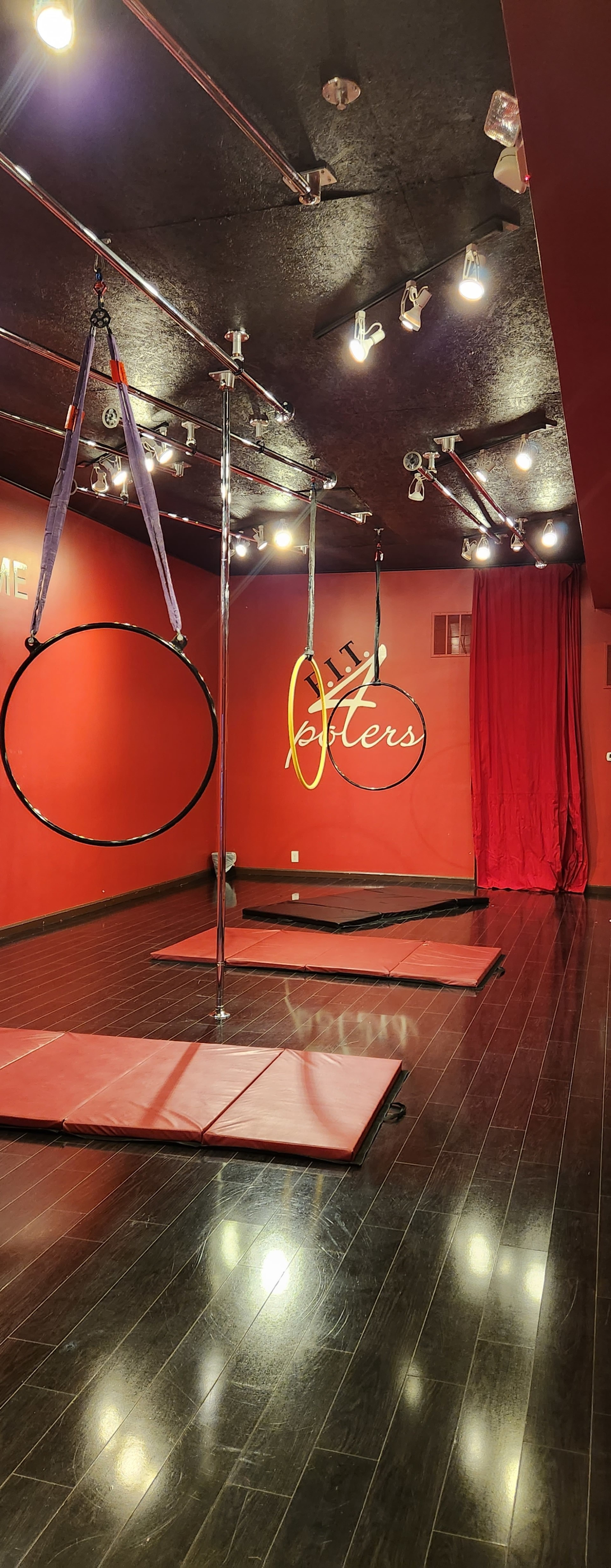 A red-walled studio featuring aerial hoops and silks suspended from the ceiling, with padded flooring beneath.