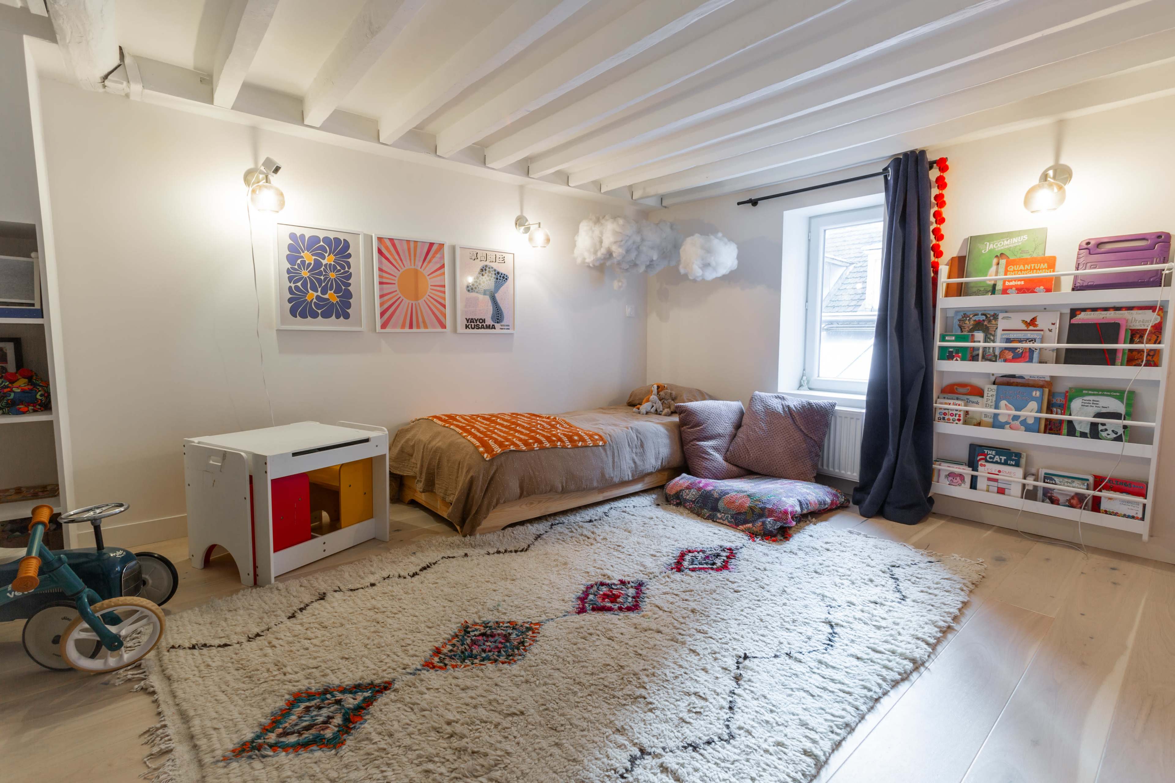A cozy children's room with a bed, bookshelves, a small table, and decorative clouds hanging from the ceiling.