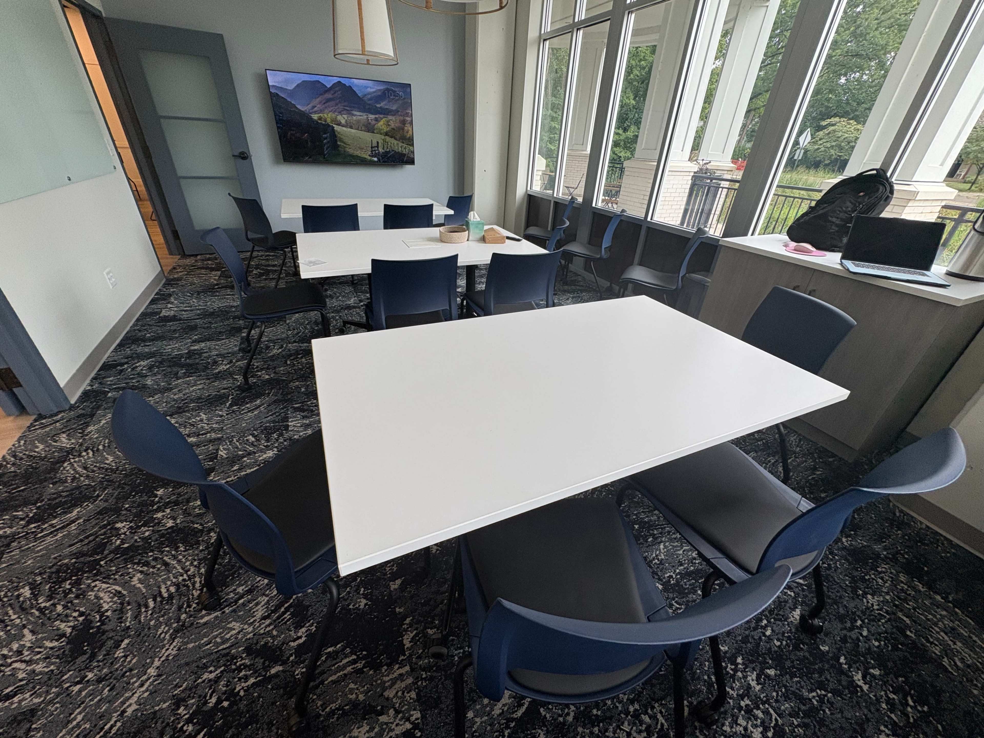 The image shows a modern conference room with a large white table surrounded by blue chairs, large windows allowing natural light, and a television mounted on the wall displaying a landscape.