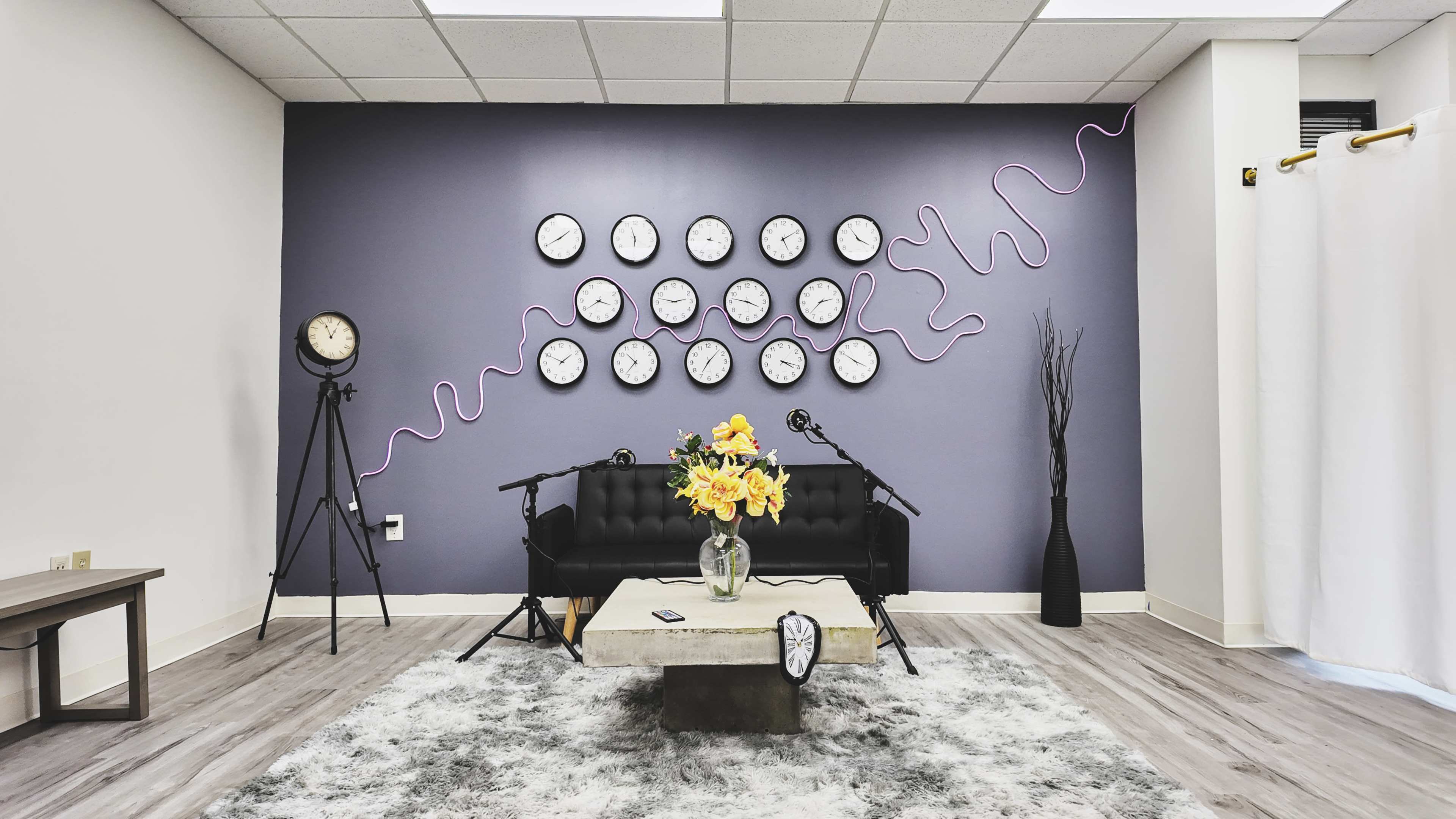 The room features a black couch with a coffee table and a large arrangement of yellow flowers, surrounded by multiple wall clocks on a purple backdrop and accent decor in a modern style.