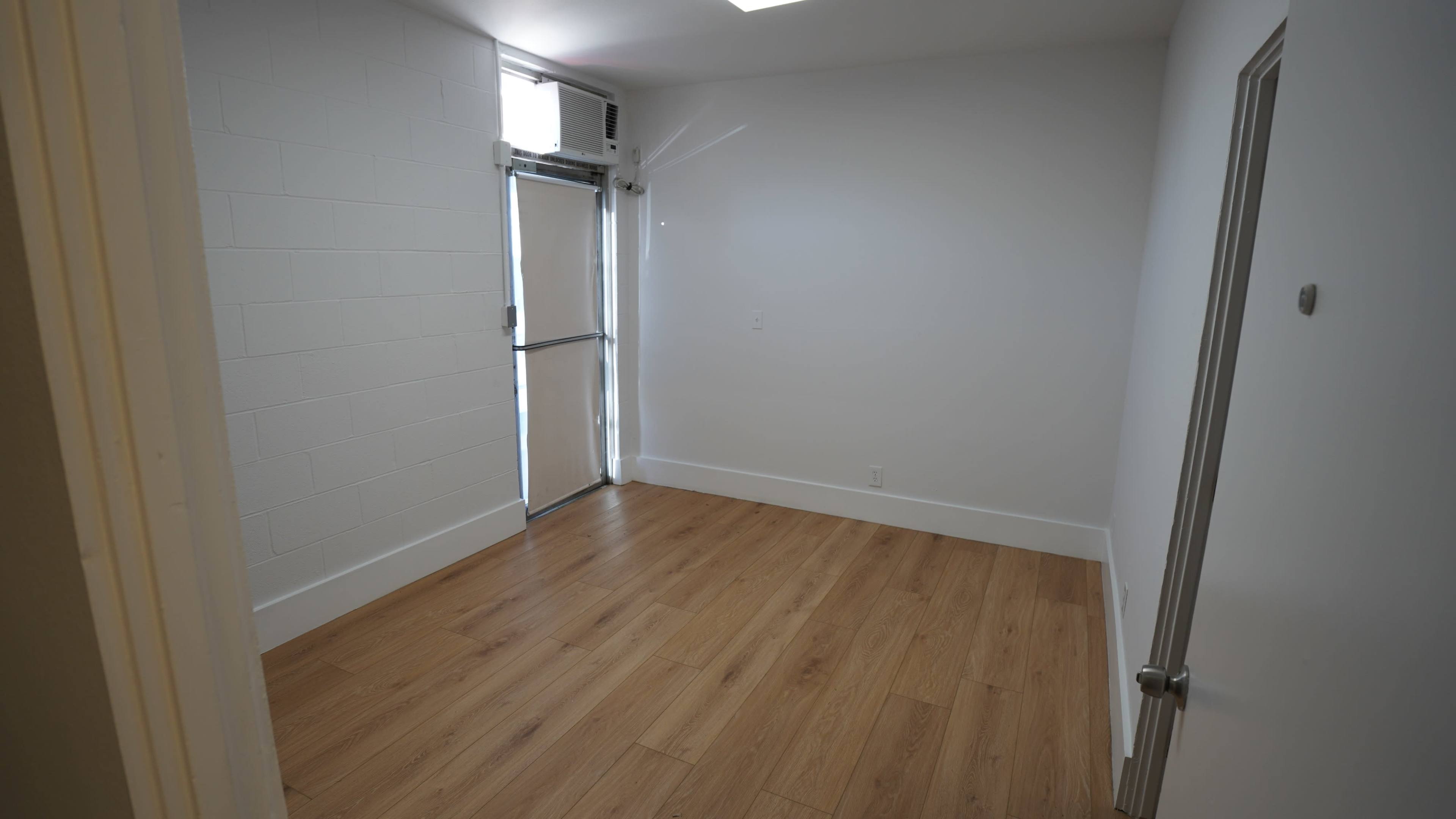 The image shows a small, empty room with wooden flooring, white walls, and a door leading to an exterior entrance.