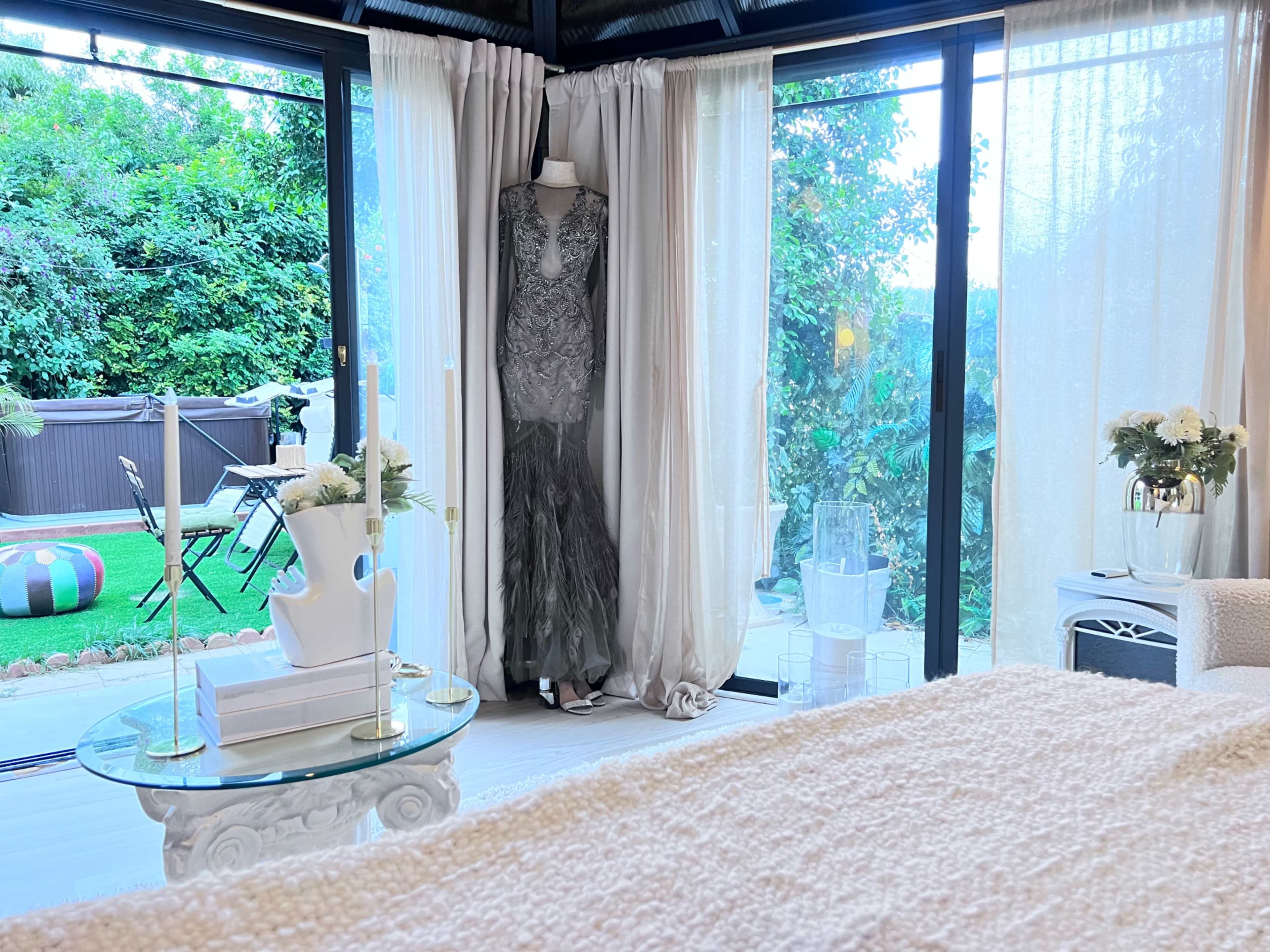 A well-lit room featuring a decorative mannequin wearing a gown, with large windows overlooking a garden and a small seating area.