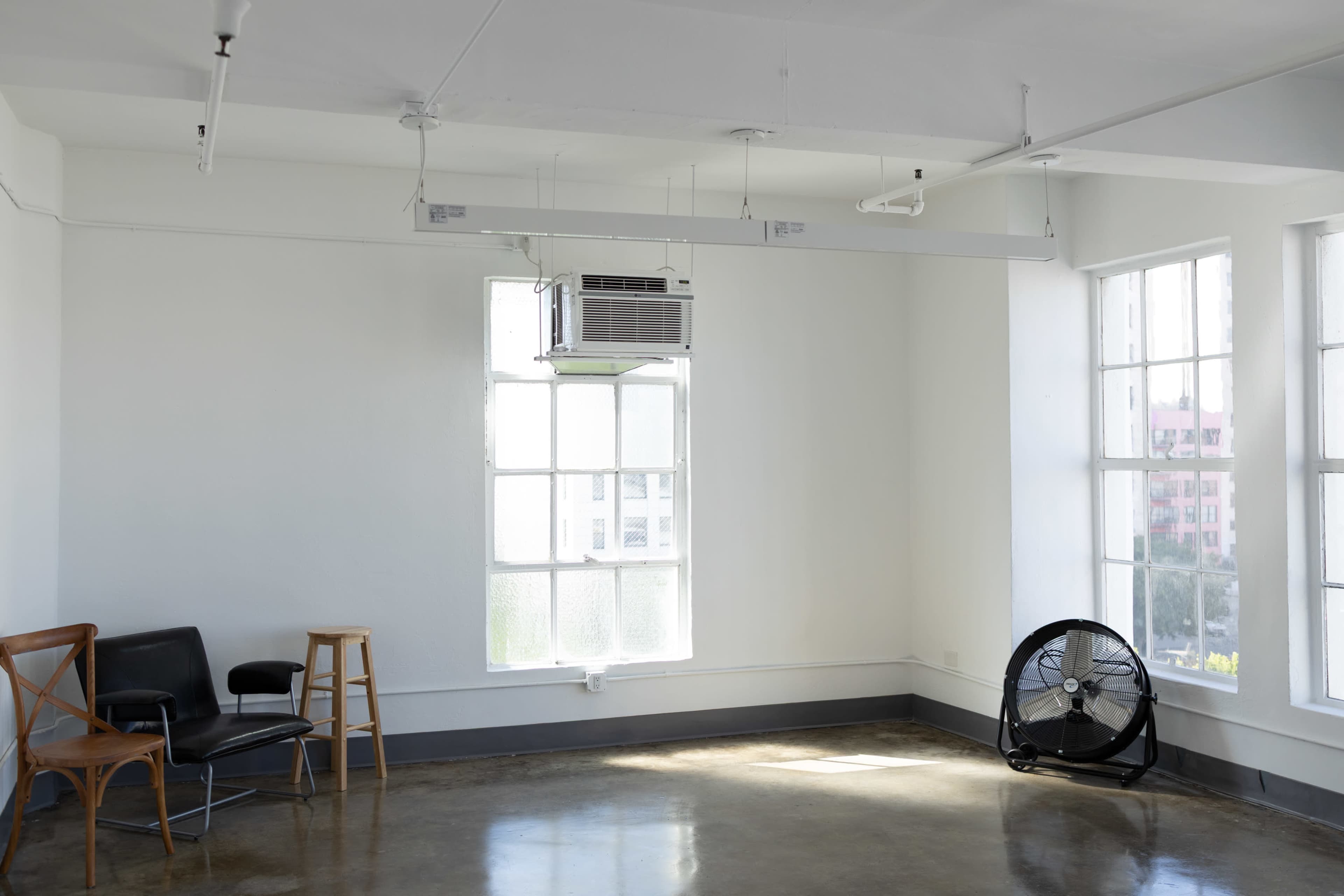 The image shows a minimalistic room with large windows, a wall-mounted air conditioner, a black chair, a wooden stool, and a floor fan.