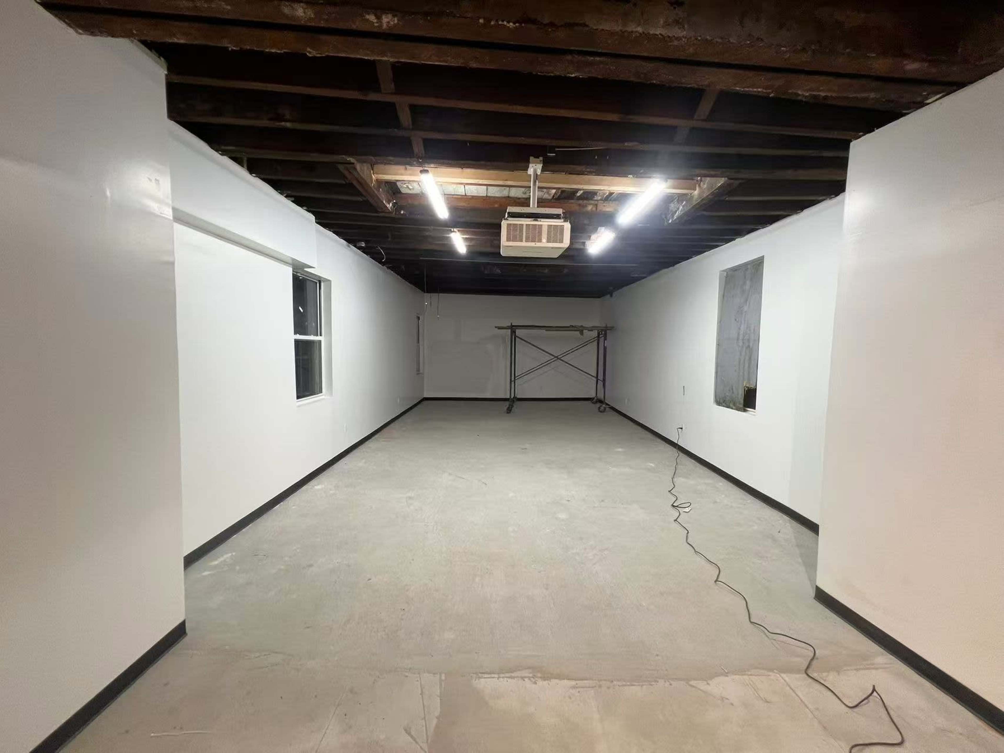 The image shows a spacious, unfinished room with exposed beams on the ceiling, white walls, and a concrete floor, along with a window and a section of scaffolding.