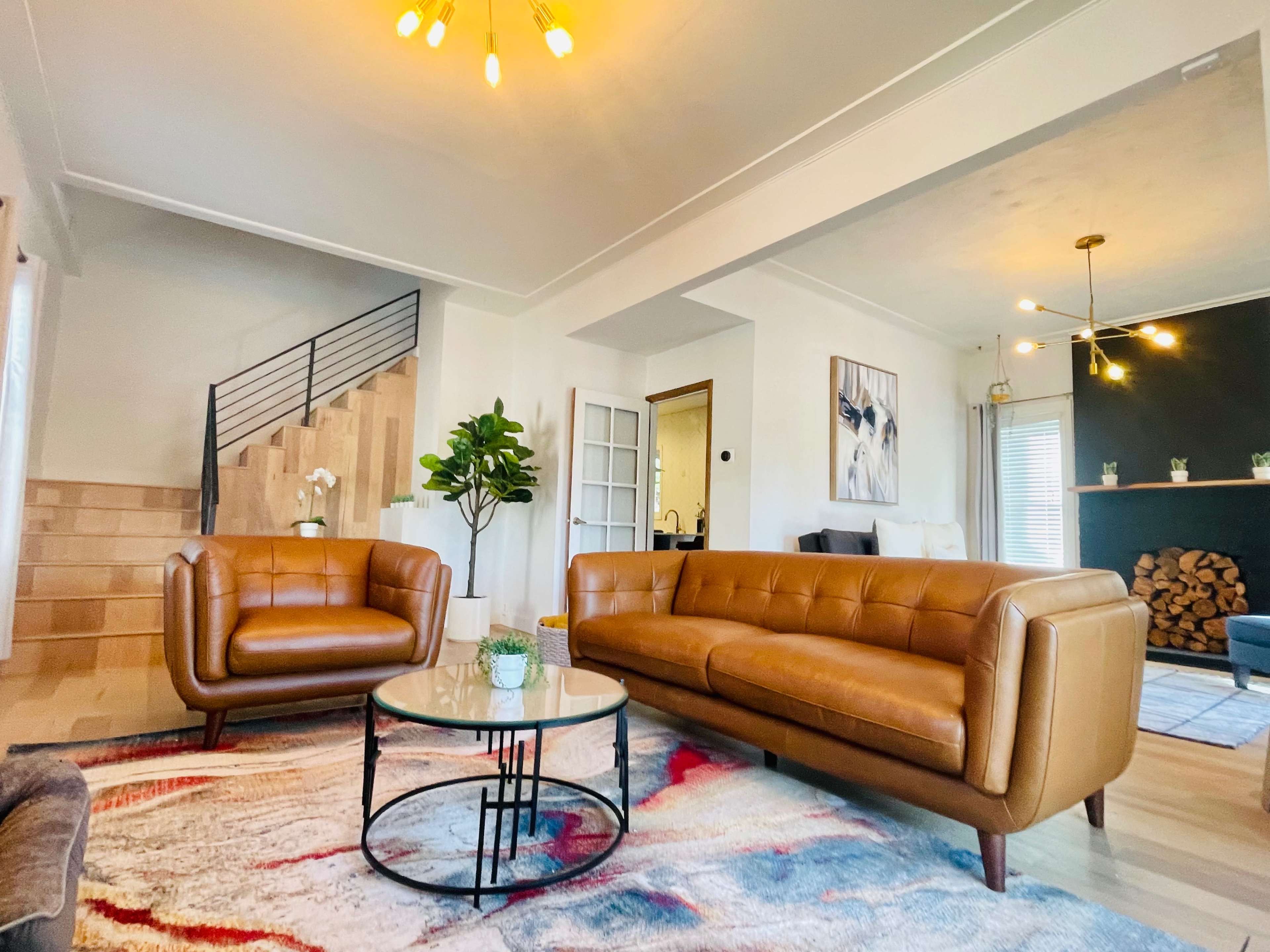 A living room features a brown leather sofa and loveseat around a circular glass coffee table on a patterned rug, with a staircase in the background.