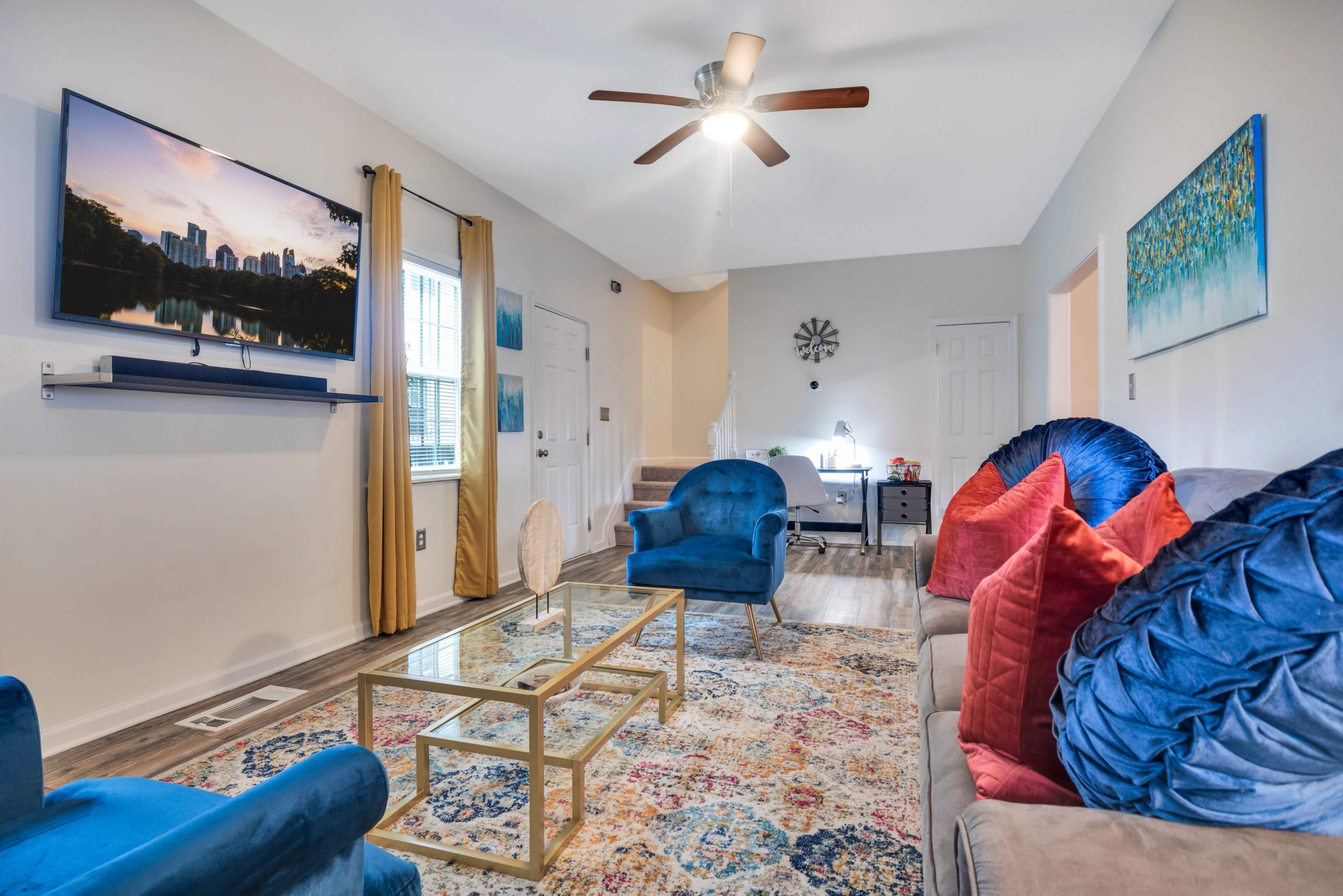 The living room features a television mounted on the wall, a glass coffee table, and blue and red upholstered seating arranged around a patterned area rug.