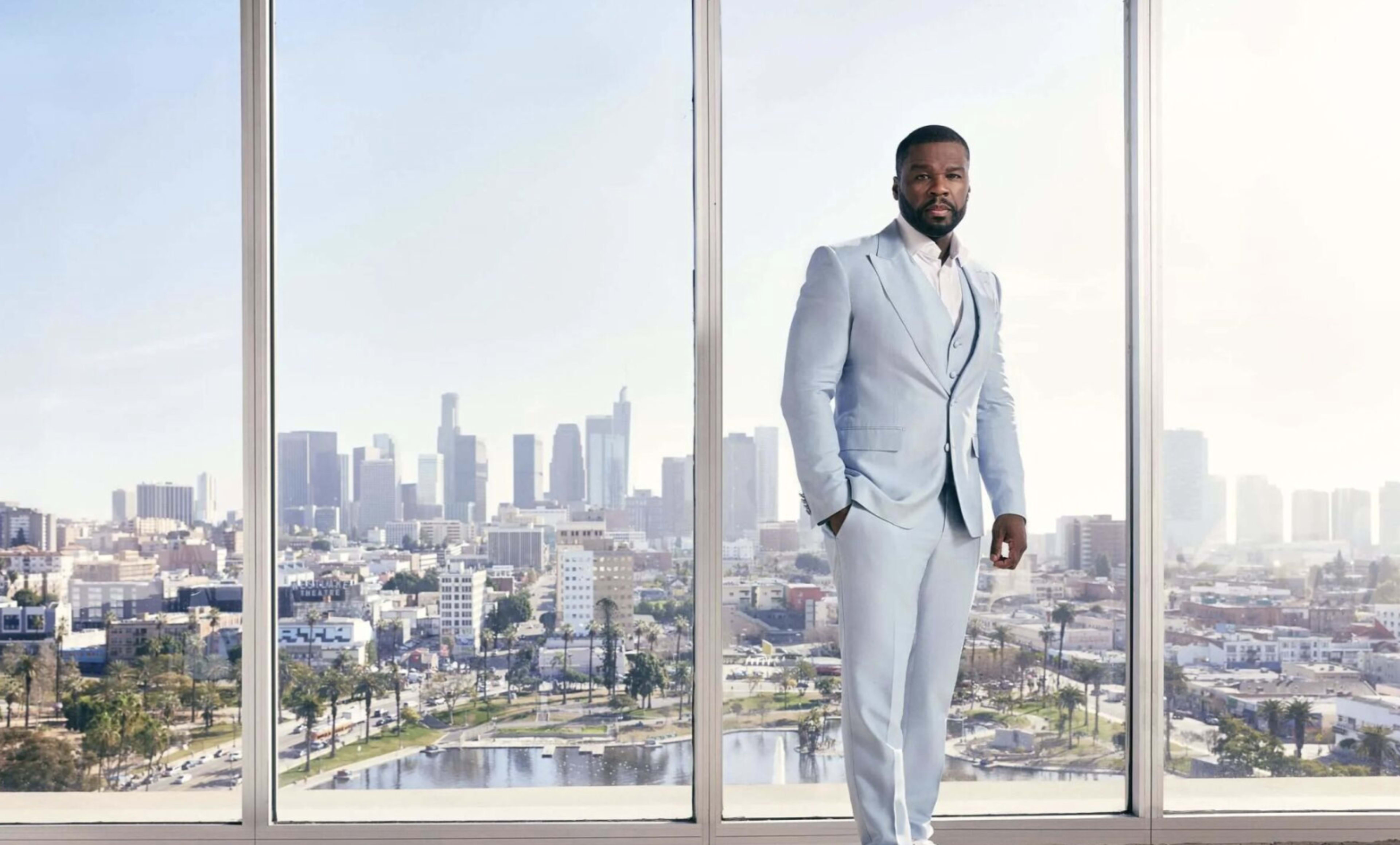 A man in a light blue suit stands in front of a large window with a view of a city skyline.