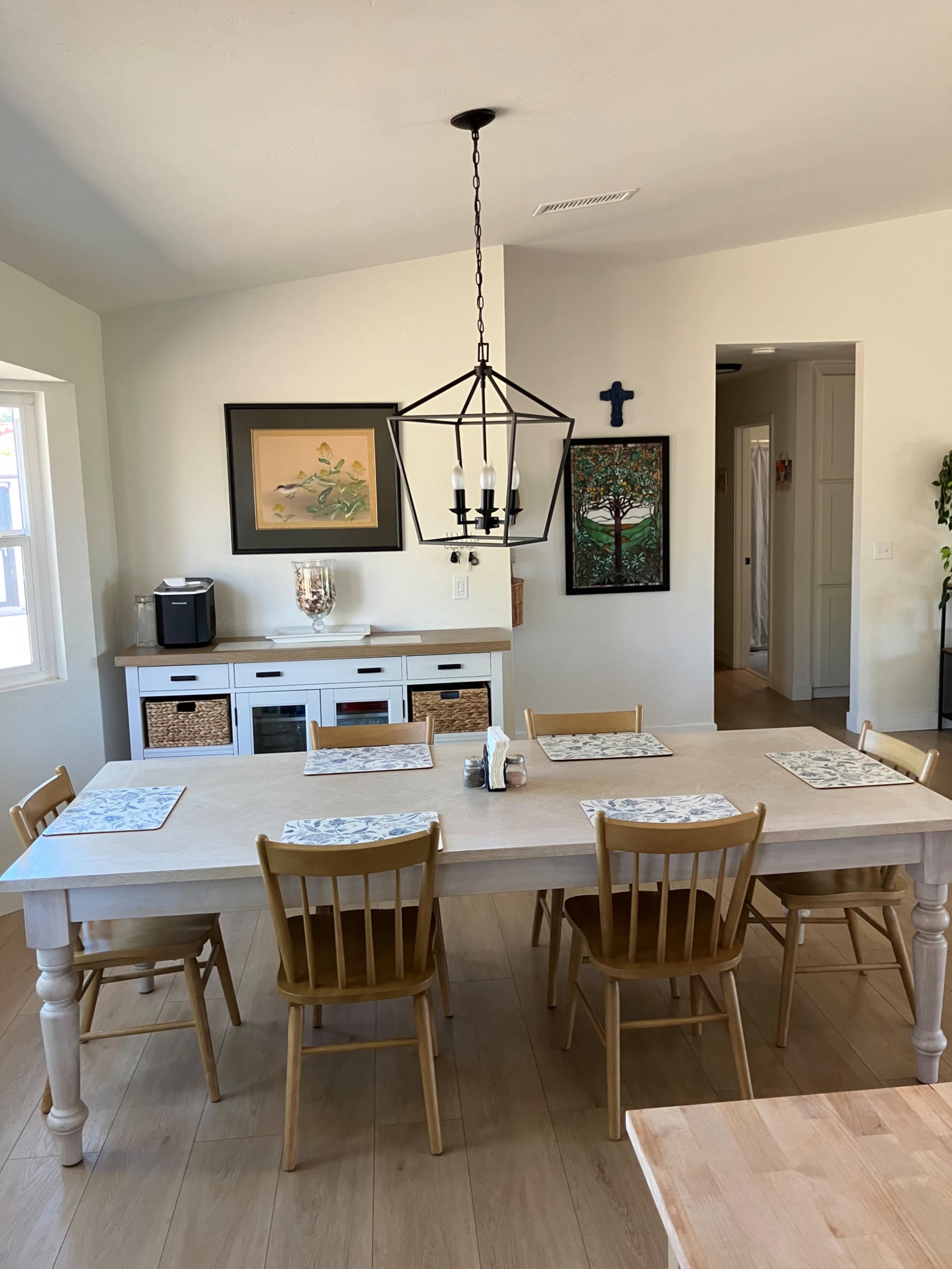 The image shows a bright dining area featuring a large wooden table surrounded by six chairs, with artwork and a cross hanging on the walls.