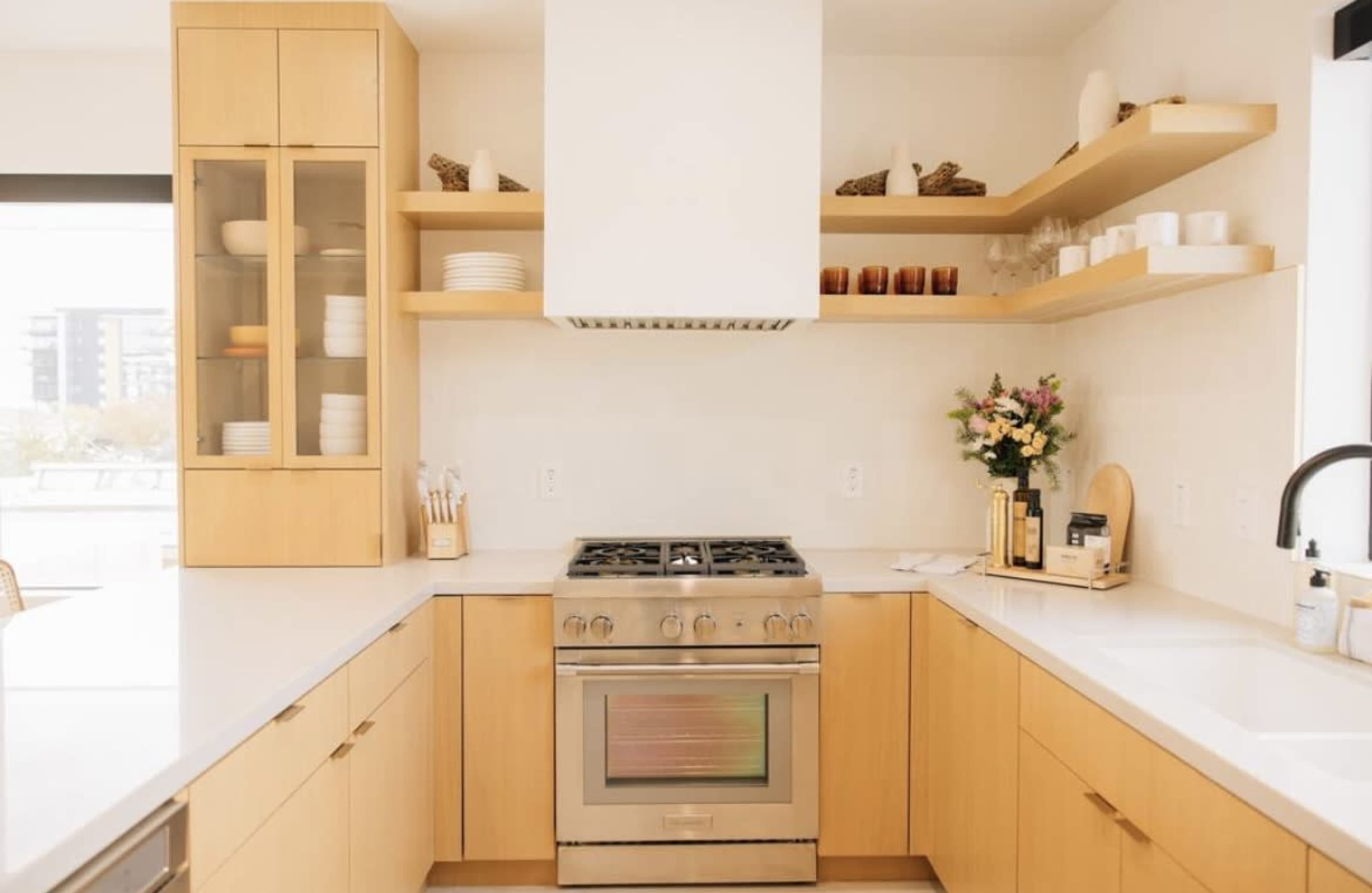 A modern kitchen features light wood cabinets, a stainless steel stove, and open shelves displaying dishes and decorative items.