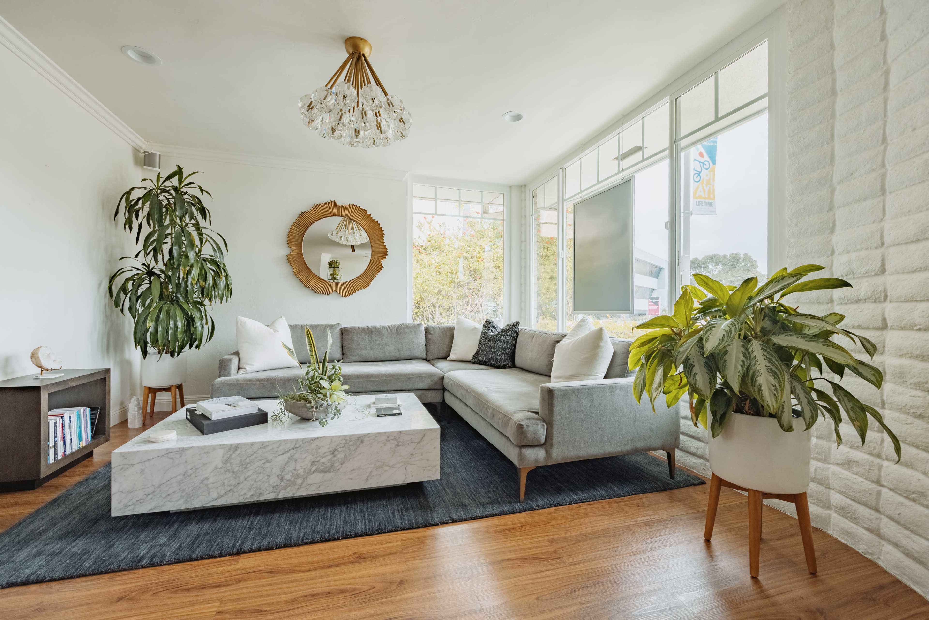 The image shows a modern living room featuring a gray sectional sofa, a marble coffee table, a large round mirror, and potted plants, with ample natural light coming through large windows.