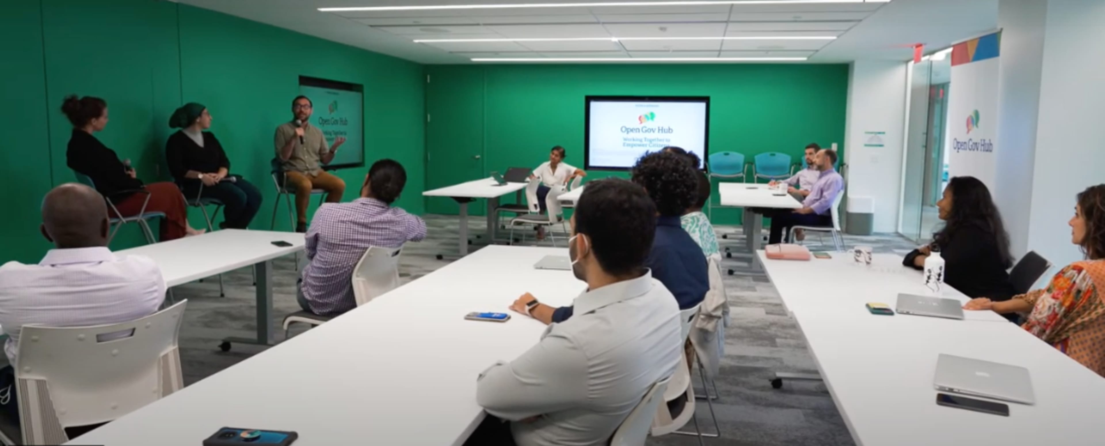 A group of individuals is seated around a conference room with green walls, engaging in a discussion while a presentation is displayed on a screen.