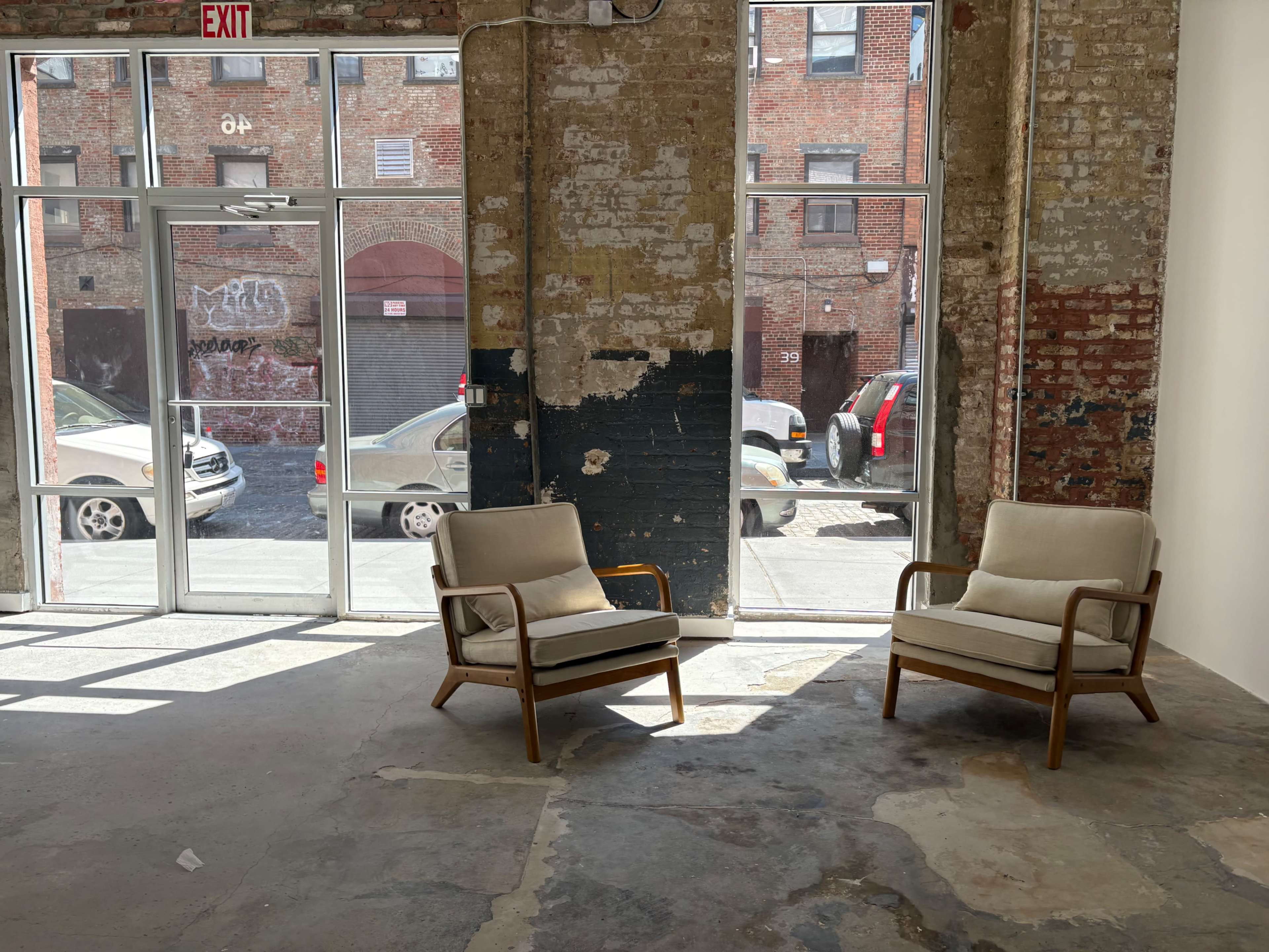 The image shows two wooden chairs with light cushions positioned in a spacious, industrial-style room featuring exposed brick walls and large windows that reveal a view of a street with parked cars.