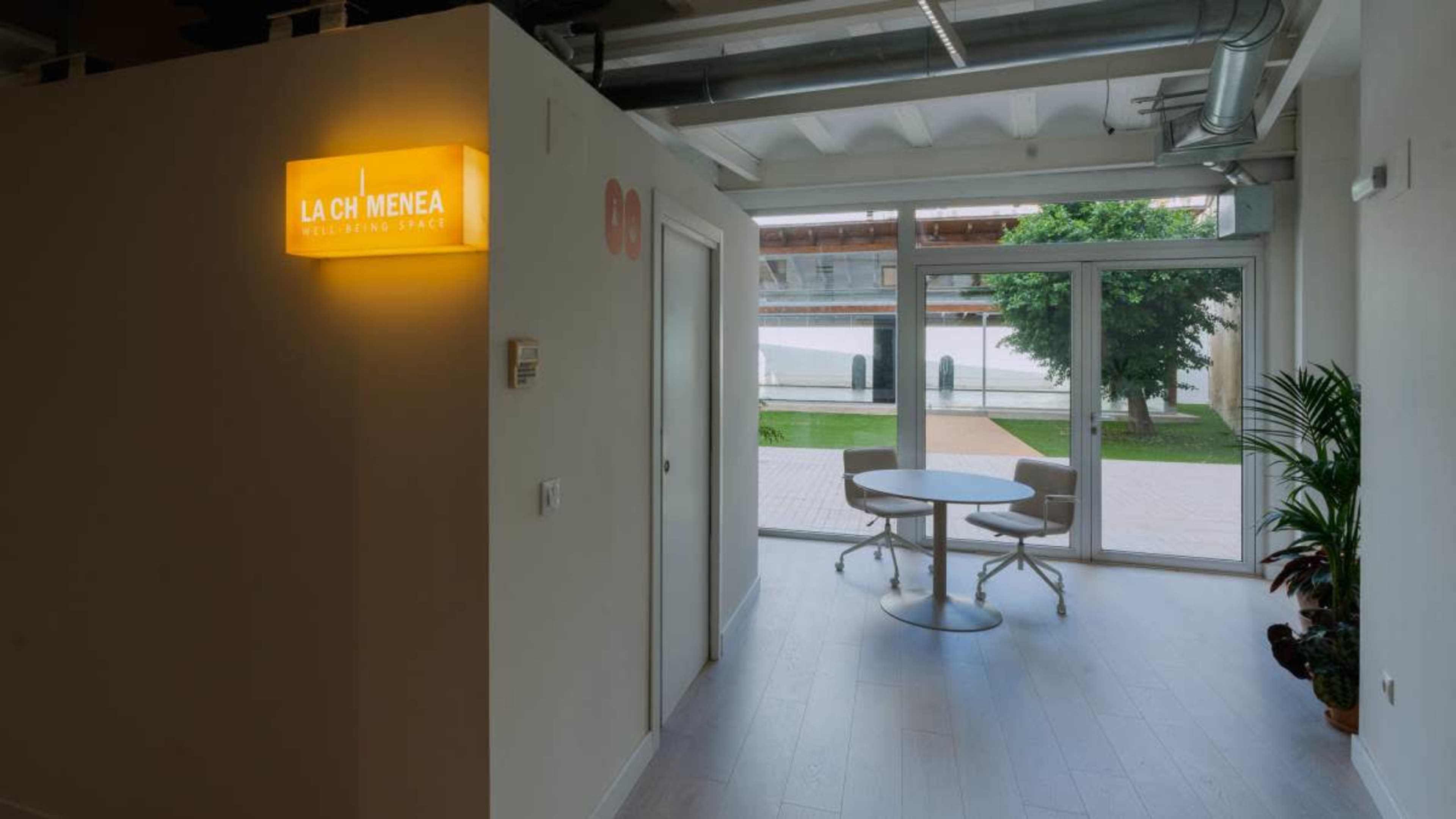 The image shows a modern interior space with a white wall displaying a yellow sign that reads "LA CHIMENEA," and a small round table set near a large window overlooking an outdoor area with greenery.