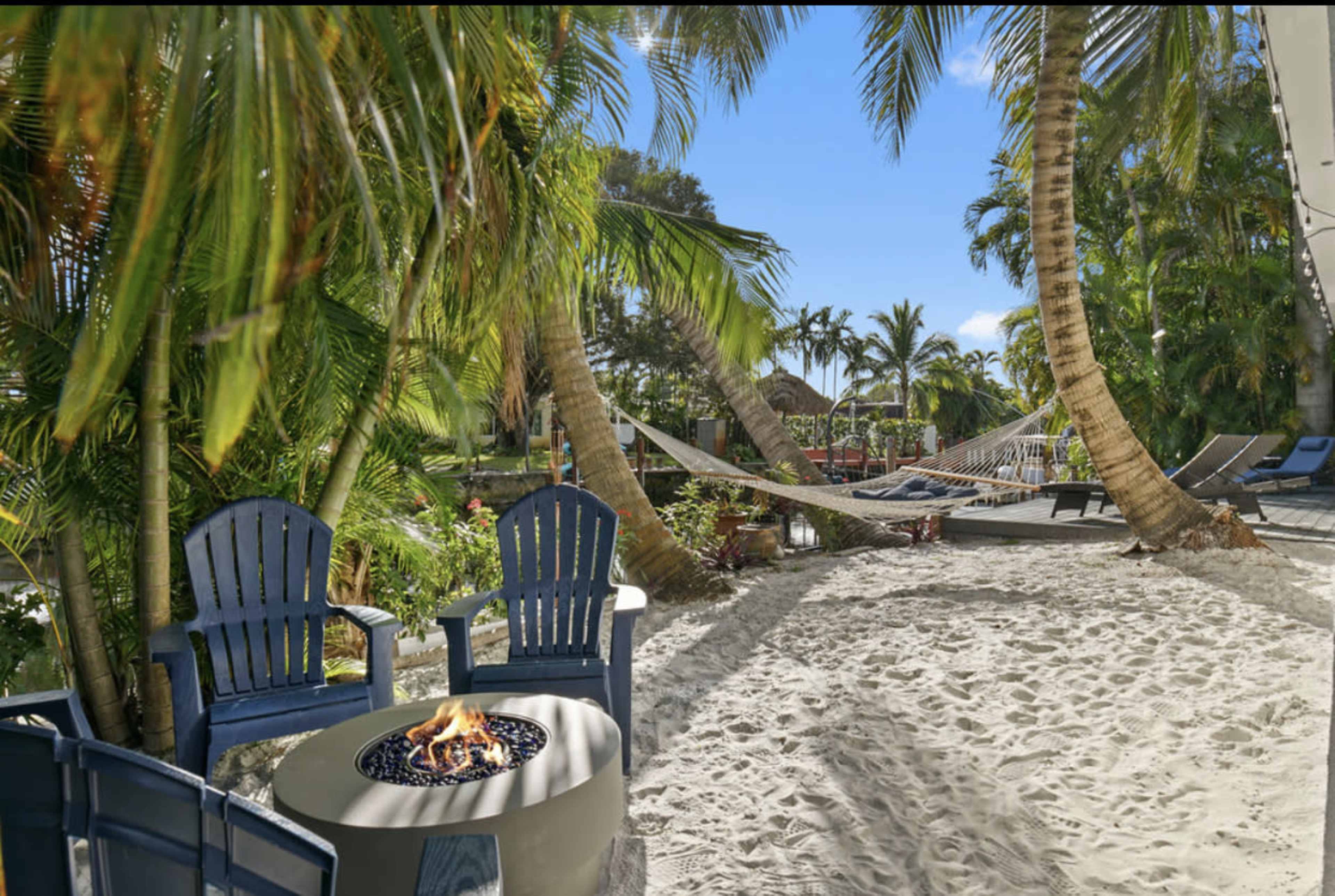 A cozy seating area with blue chairs and a fire pit is surrounded by palm trees and sandy ground, with hammocks and lounge chairs in the background.