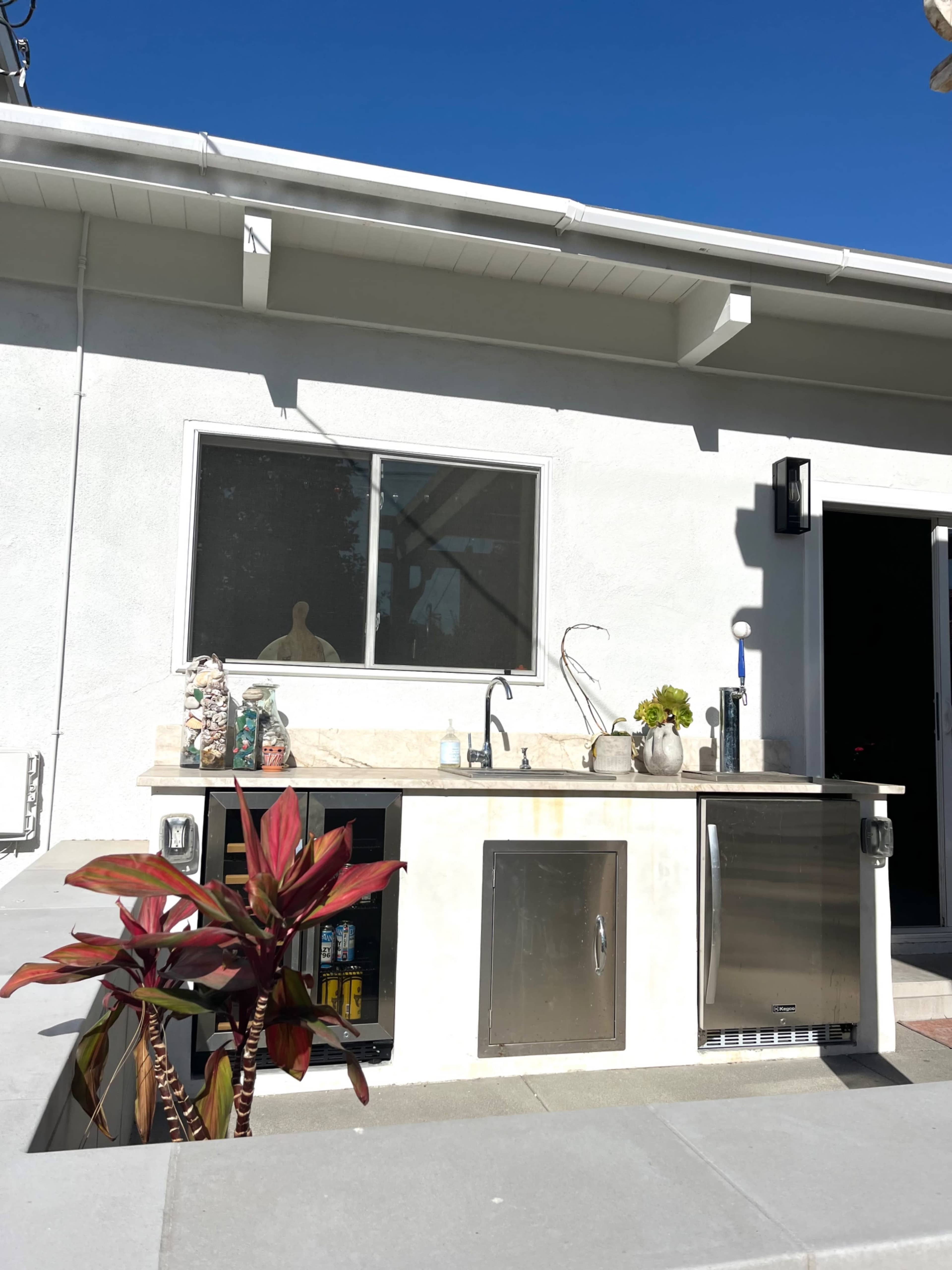 The image shows an outdoor kitchen area with a countertop, sink, refrigerator, and a potted plant in front.