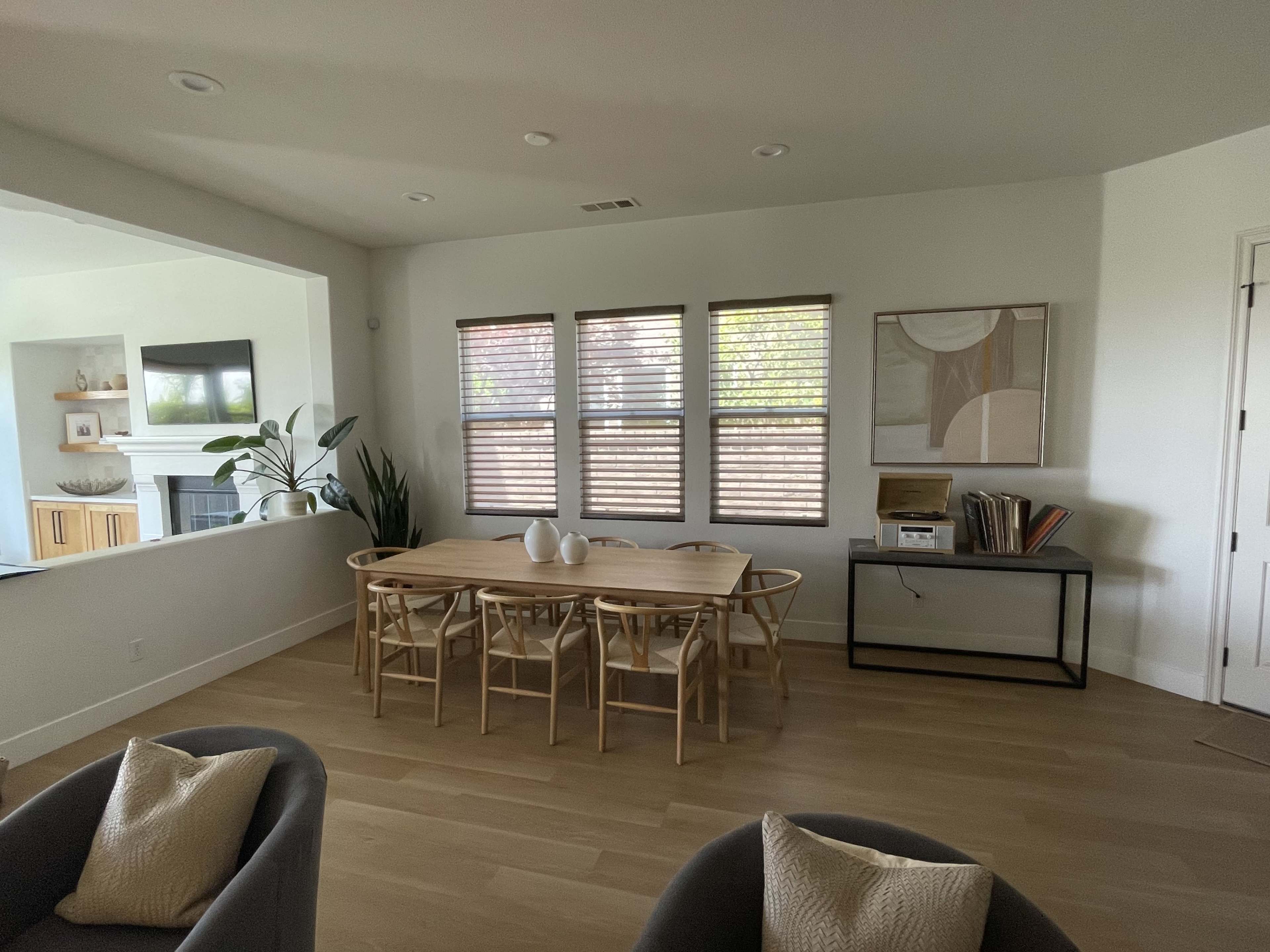 A modern dining area features a wooden table with chairs, positioned near large windows with blinds, beside a console table and artwork on the wall.