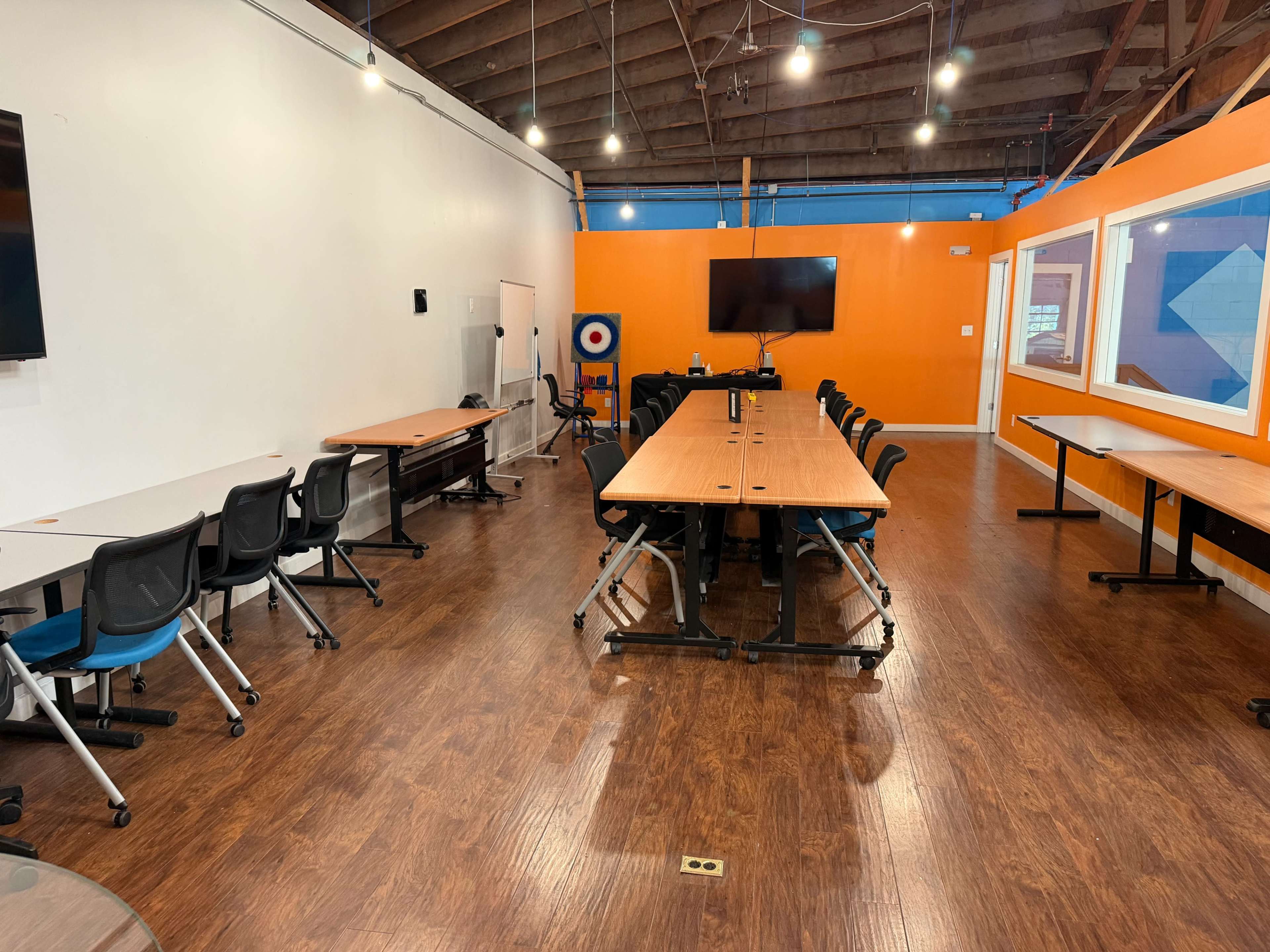 The image shows a spacious, well-lit meeting room with several long tables arranged for group seating and bright orange accent walls.