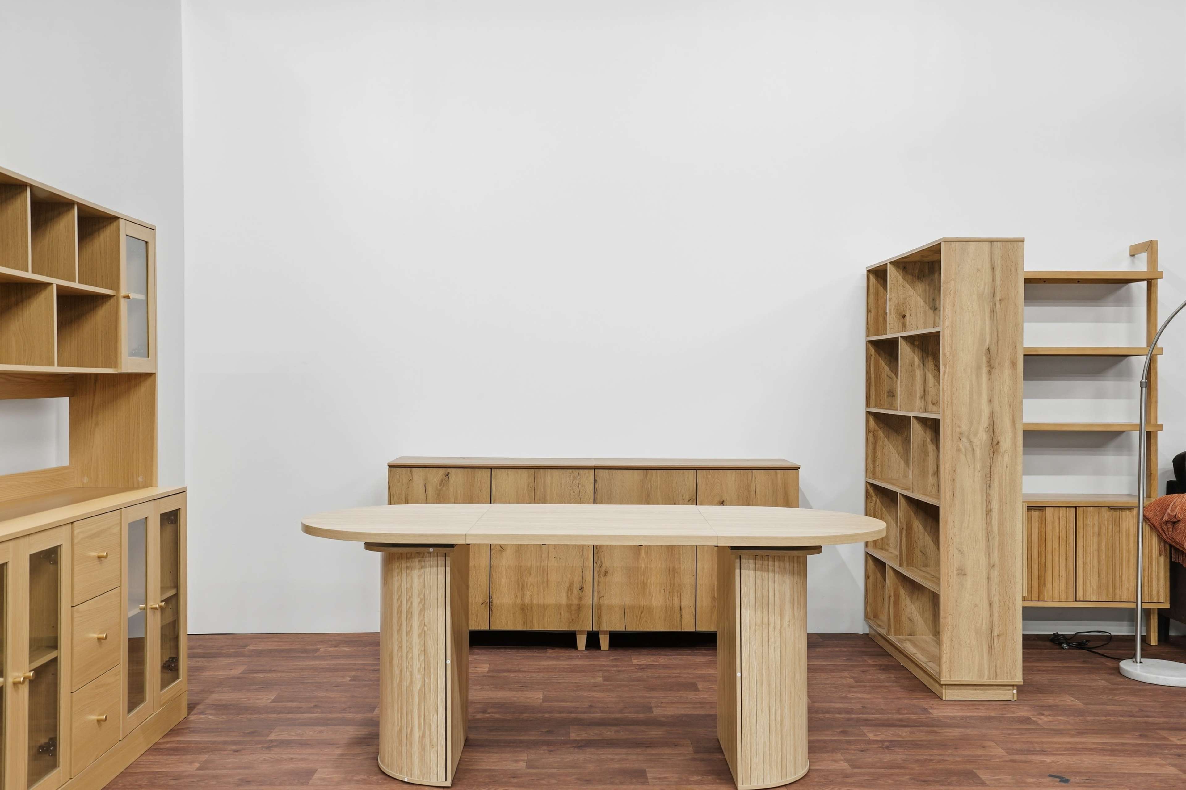 The image shows a light wood-furnished office space featuring a central curved desk, two wooden shelving units, and a side cabinet against a white wall.