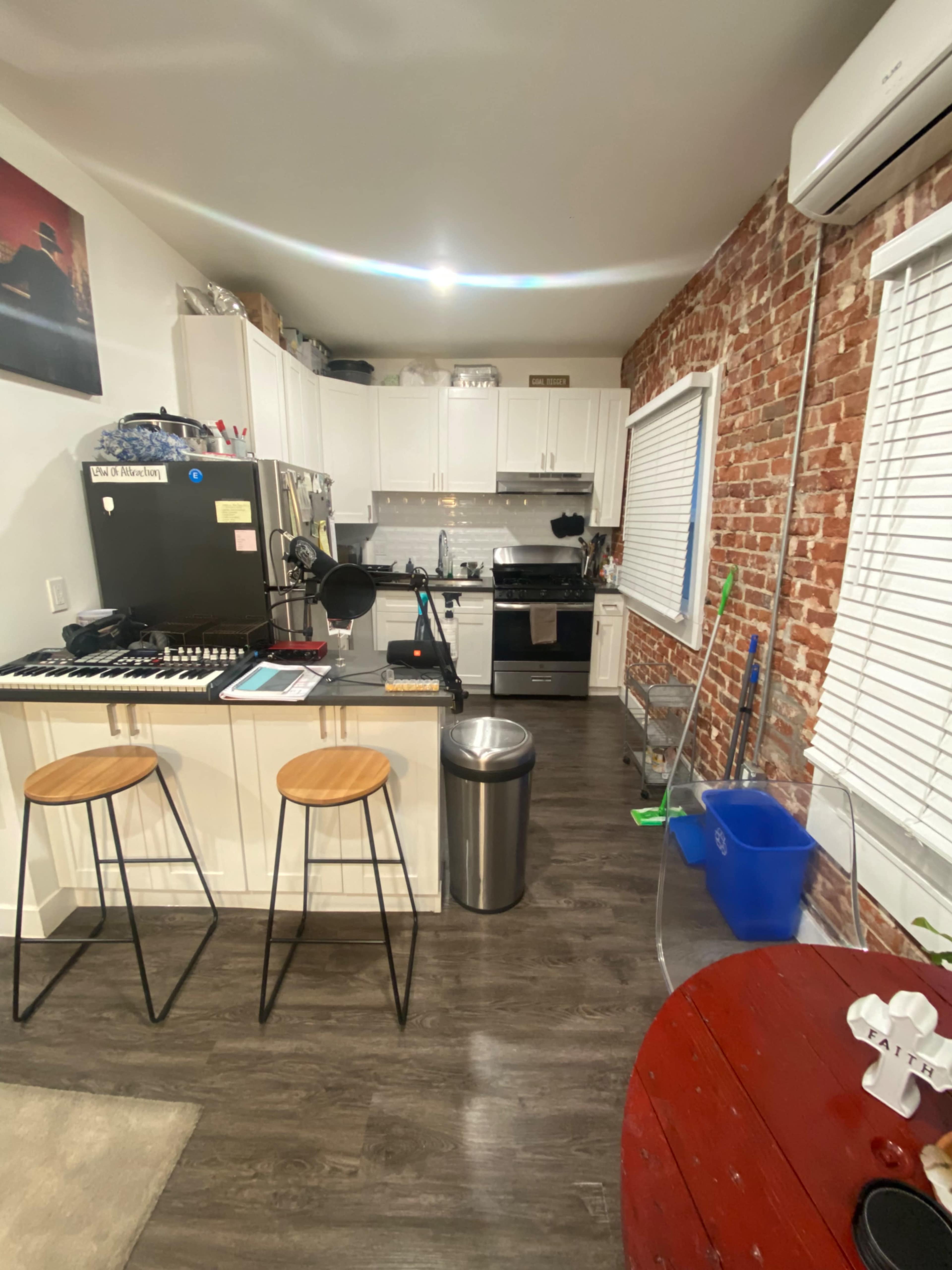 The image shows a compact kitchen area with white cabinets, a black refrigerator, a stove, and two wooden stools at a small table, featuring exposed brick walls and modern appliances.