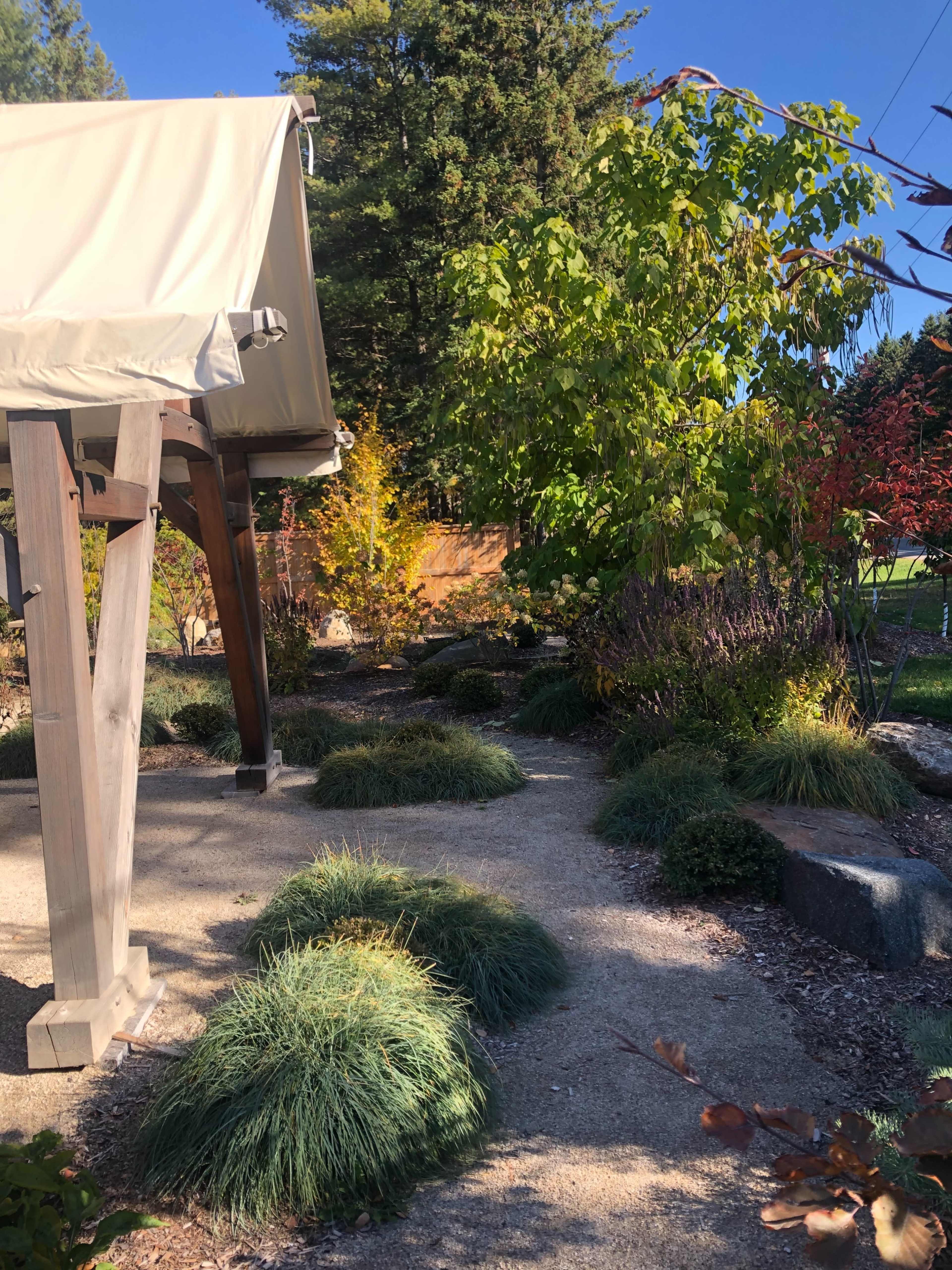 A landscaped garden features a wooden structure with a canvas roof surrounded by various plants and gravel pathways.