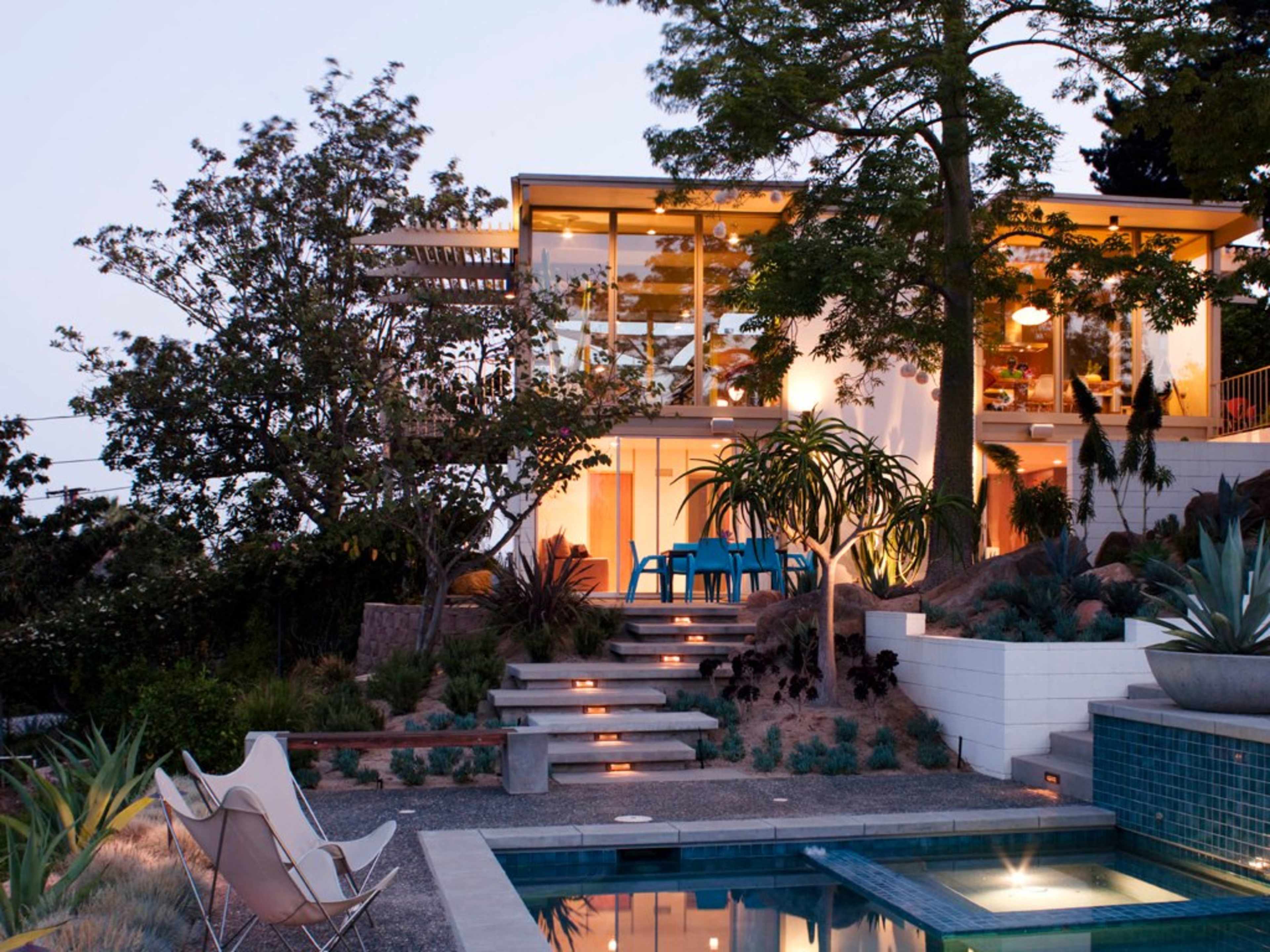 A modern house with large glass windows and a pool is surrounded by landscaped gardens and walkways leading up to it.