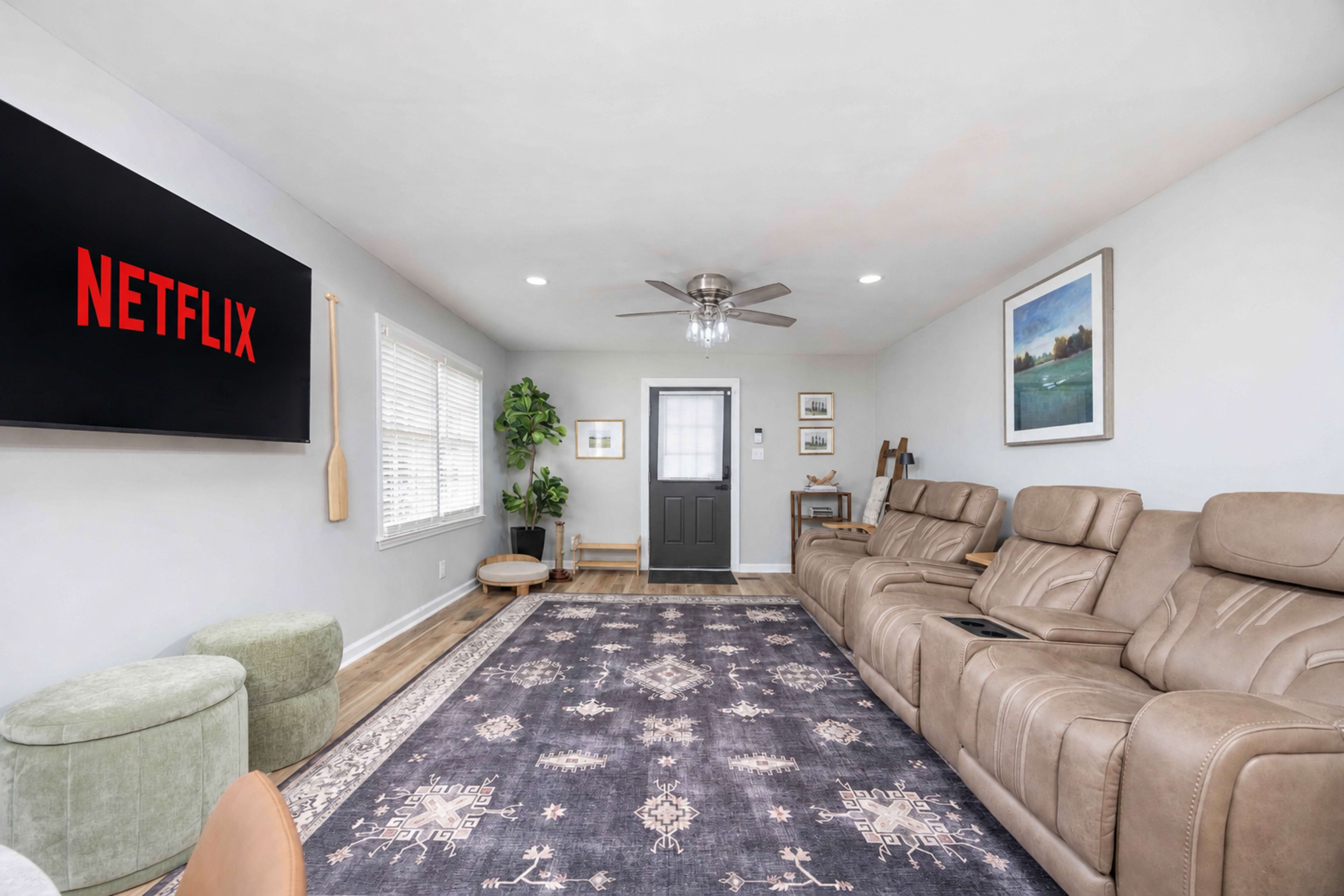 The image shows a modern living room featuring a large Netflix screen, comfortable recliner seating, a decorative rug, and a door leading outside.