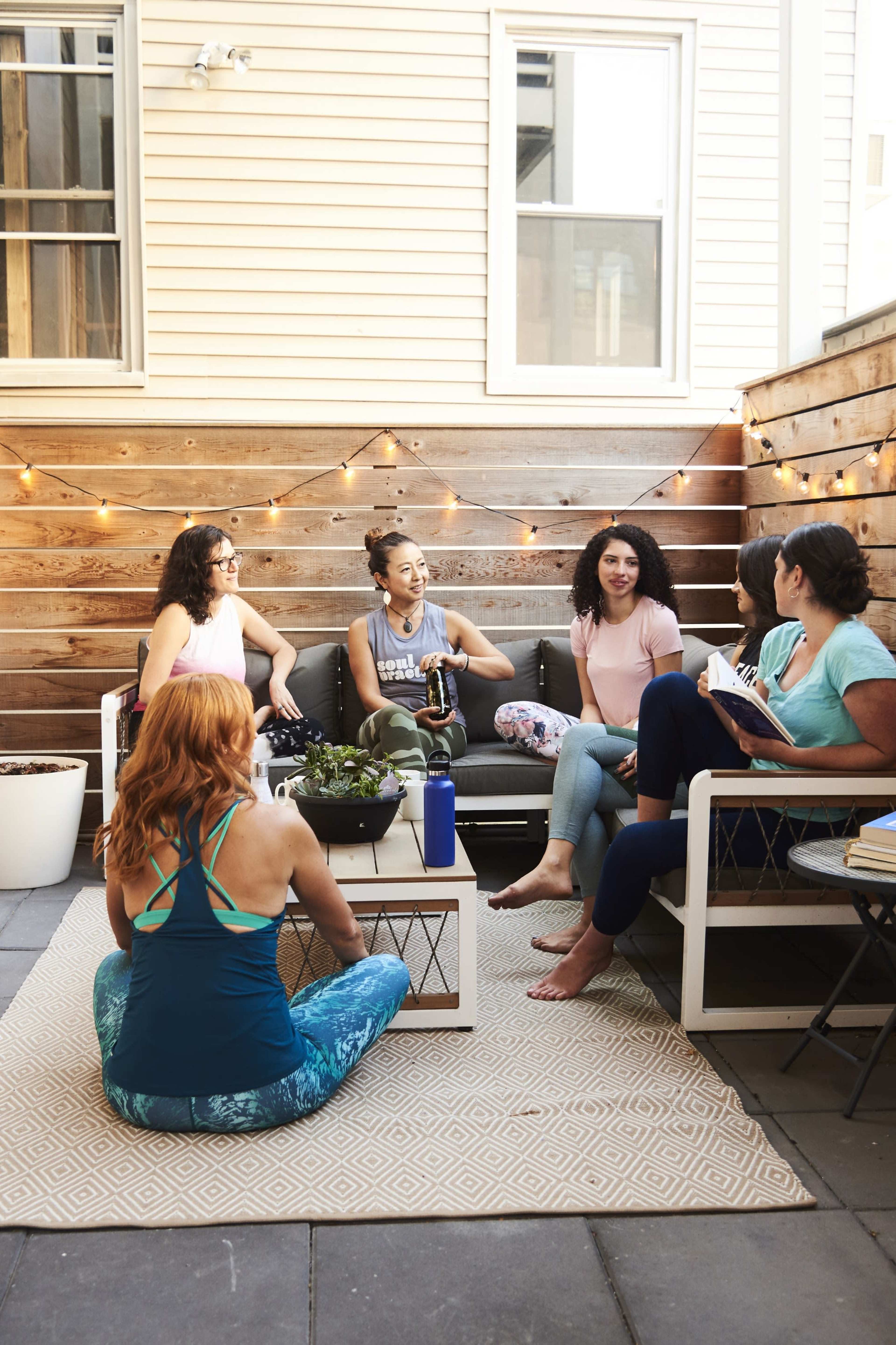A group of six women sits in a cozy outdoor seating area, chatting and enjoying each other's company, with string lights illuminating the space.