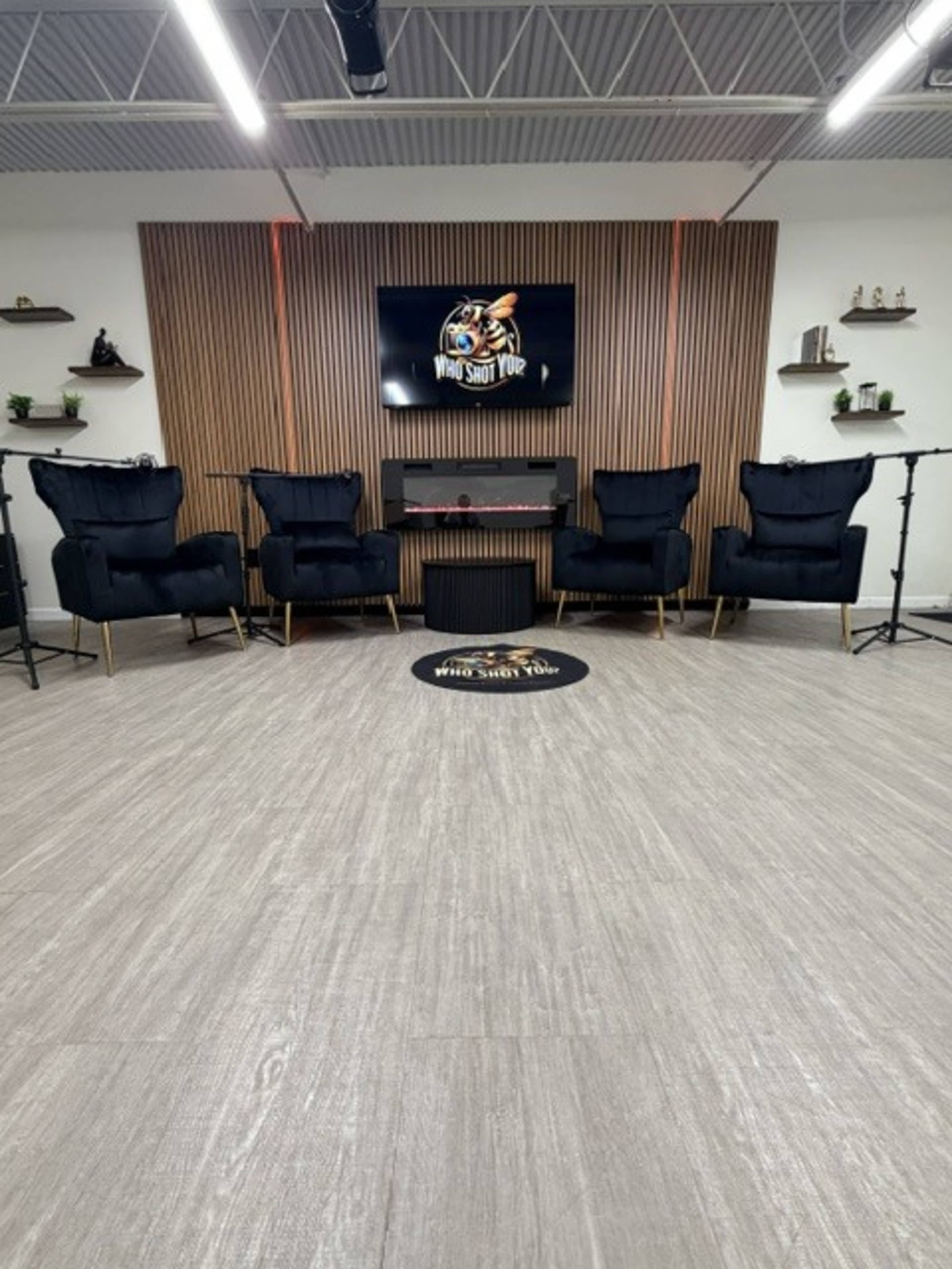 The image shows a modern room with a smooth wooden floor, four black armchairs arranged in a semi-circle, a low table, and a TV mounted on the wall displaying a logo.