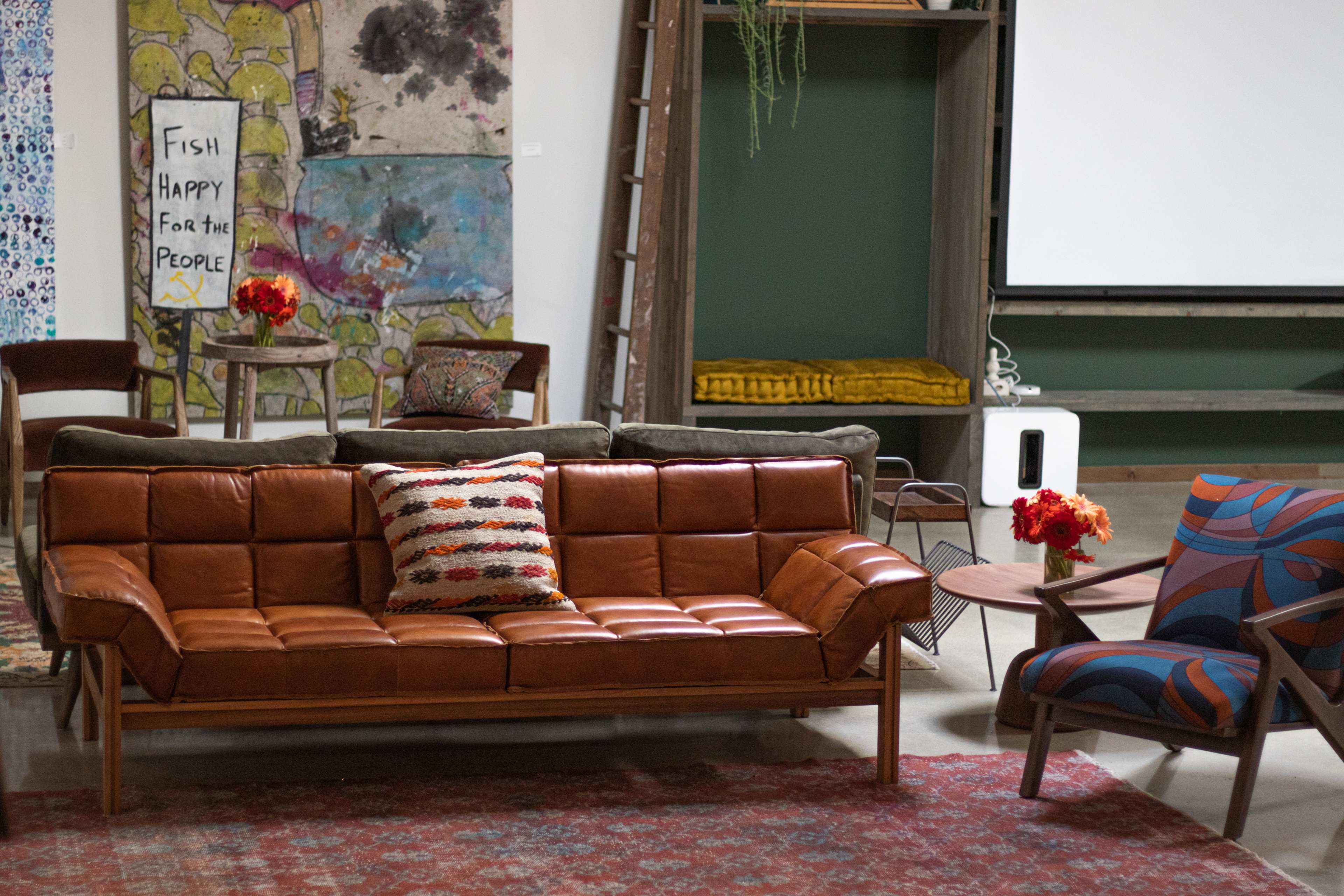 The image shows a modern interior space featuring a brown leather sofa, a patterned armchair, a wooden coffee table, and colorful floral arrangements.