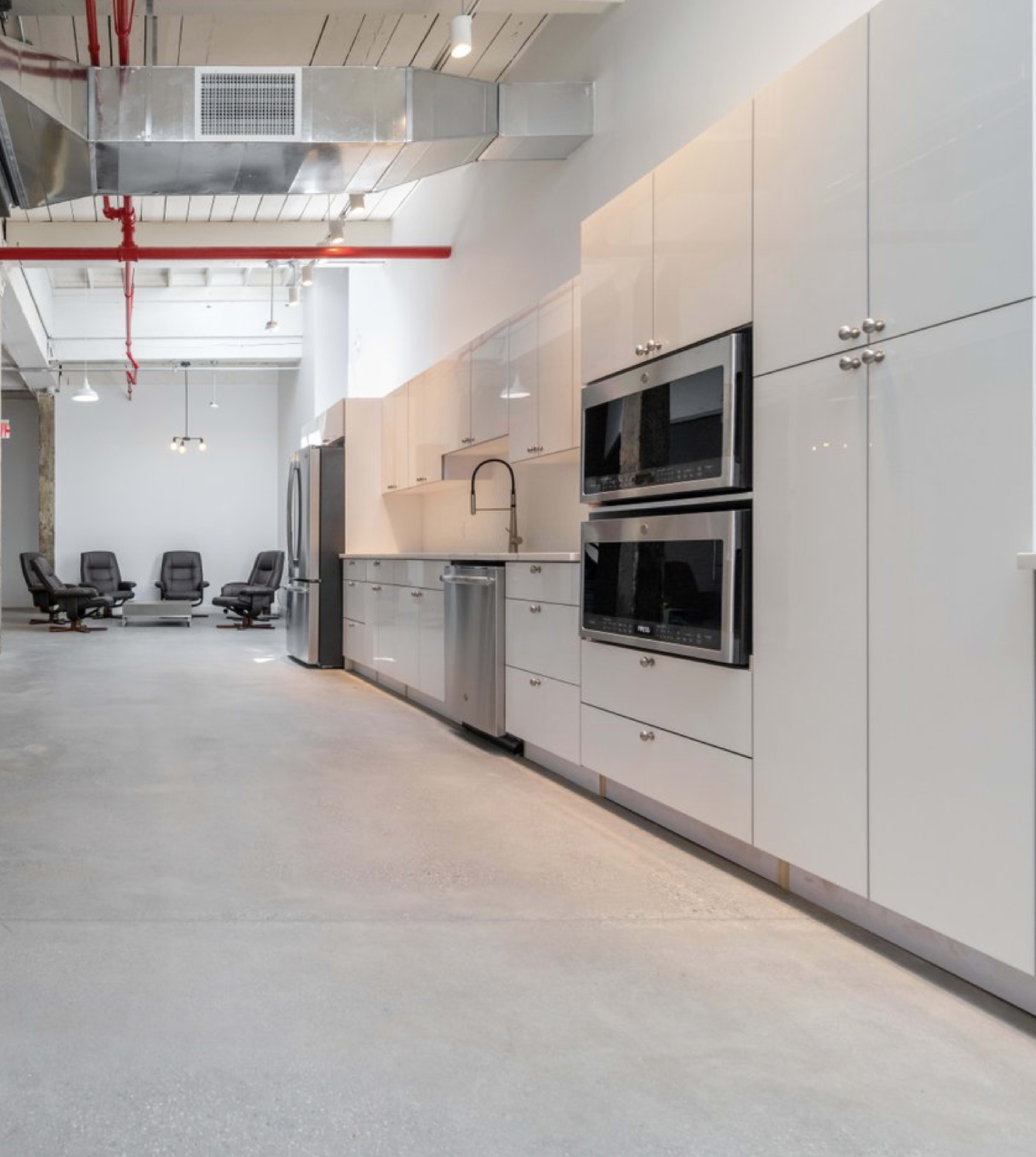 The image shows a modern kitchen area with white cabinetry, stainless steel appliances, and a polished concrete floor, leading into a seating area with black chairs in the background.