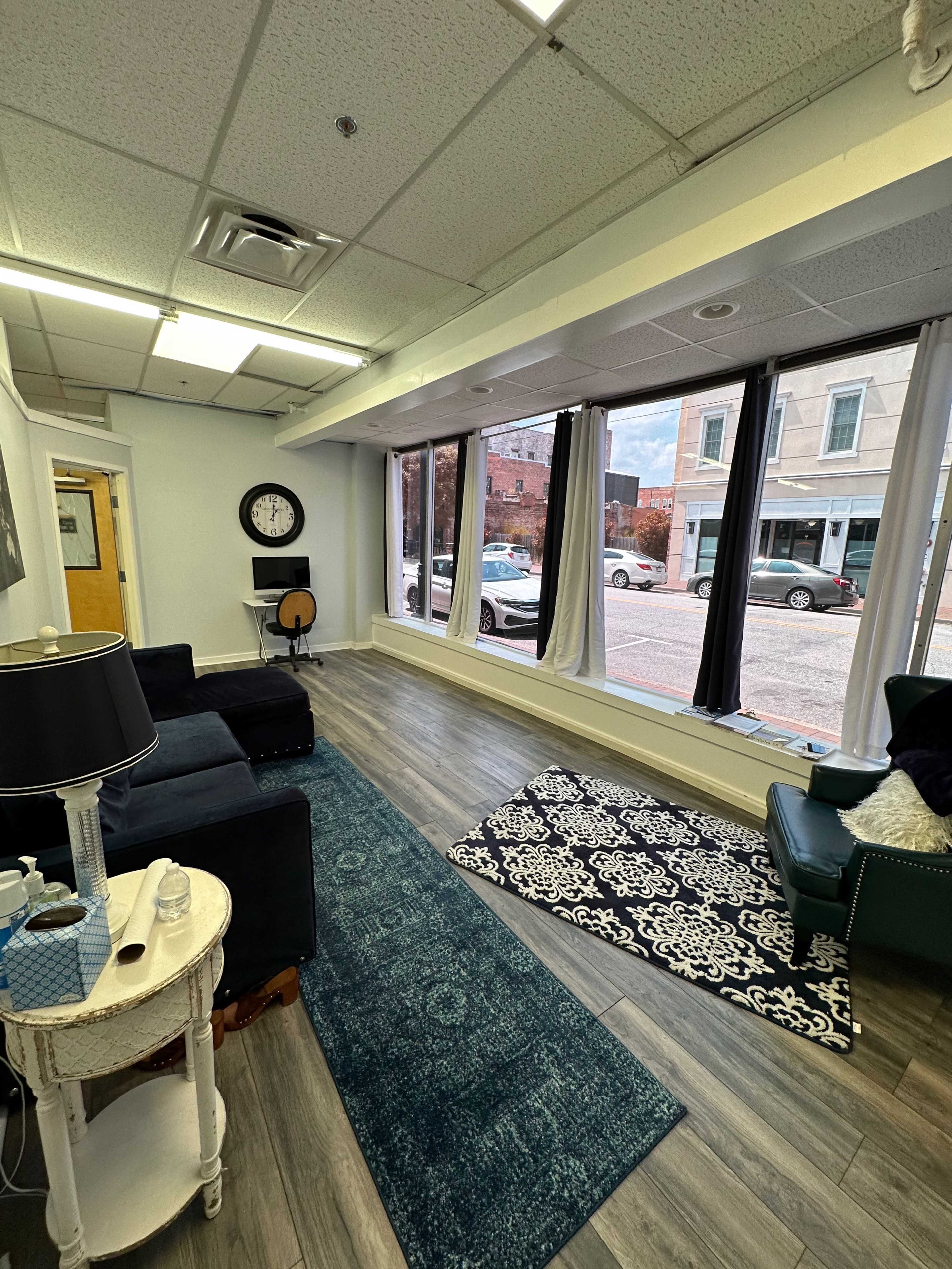 A brightly lit interior of a room with hardwood flooring, featuring two seating areas, large windows facing the street, and decorative rugs.