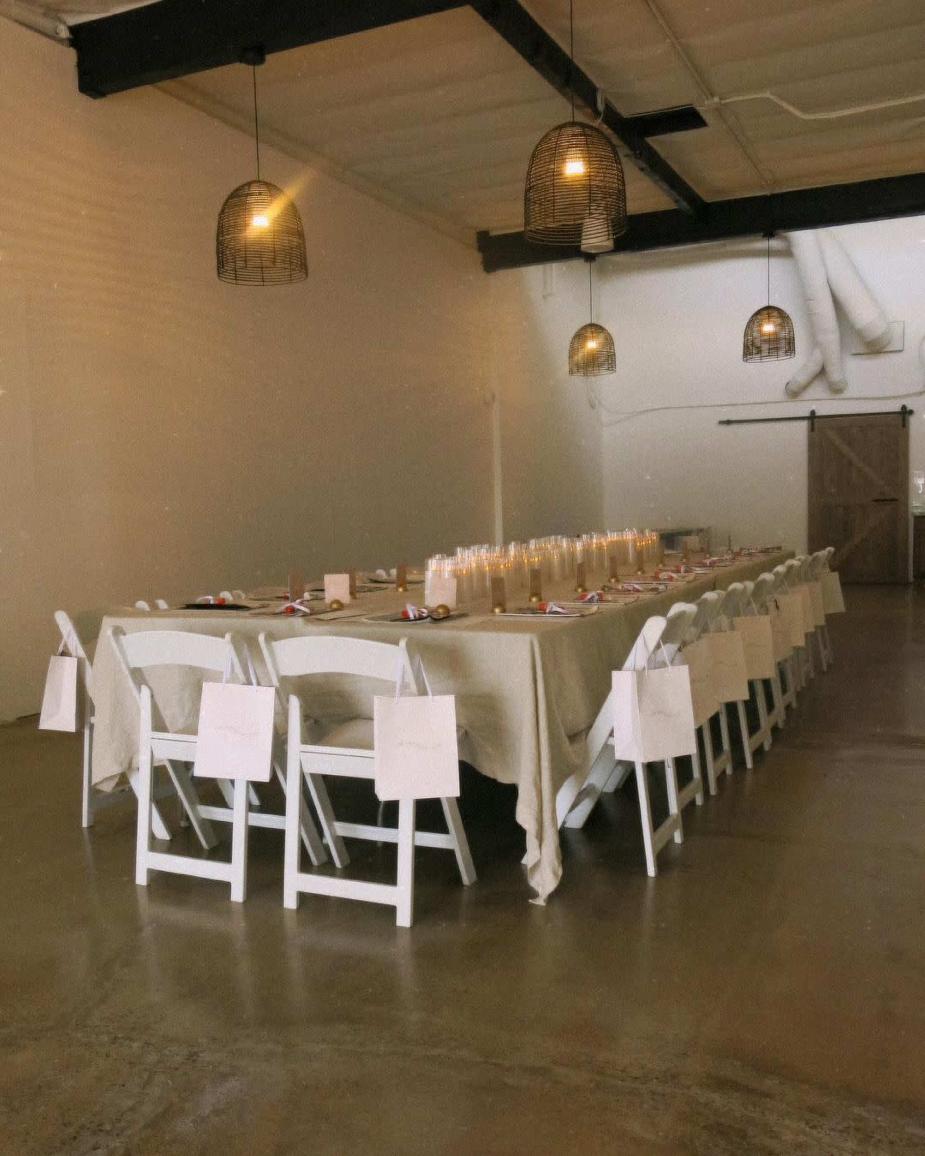 A long, folded table is set with white chairs and simple place settings under pendant lights in a spacious room.
