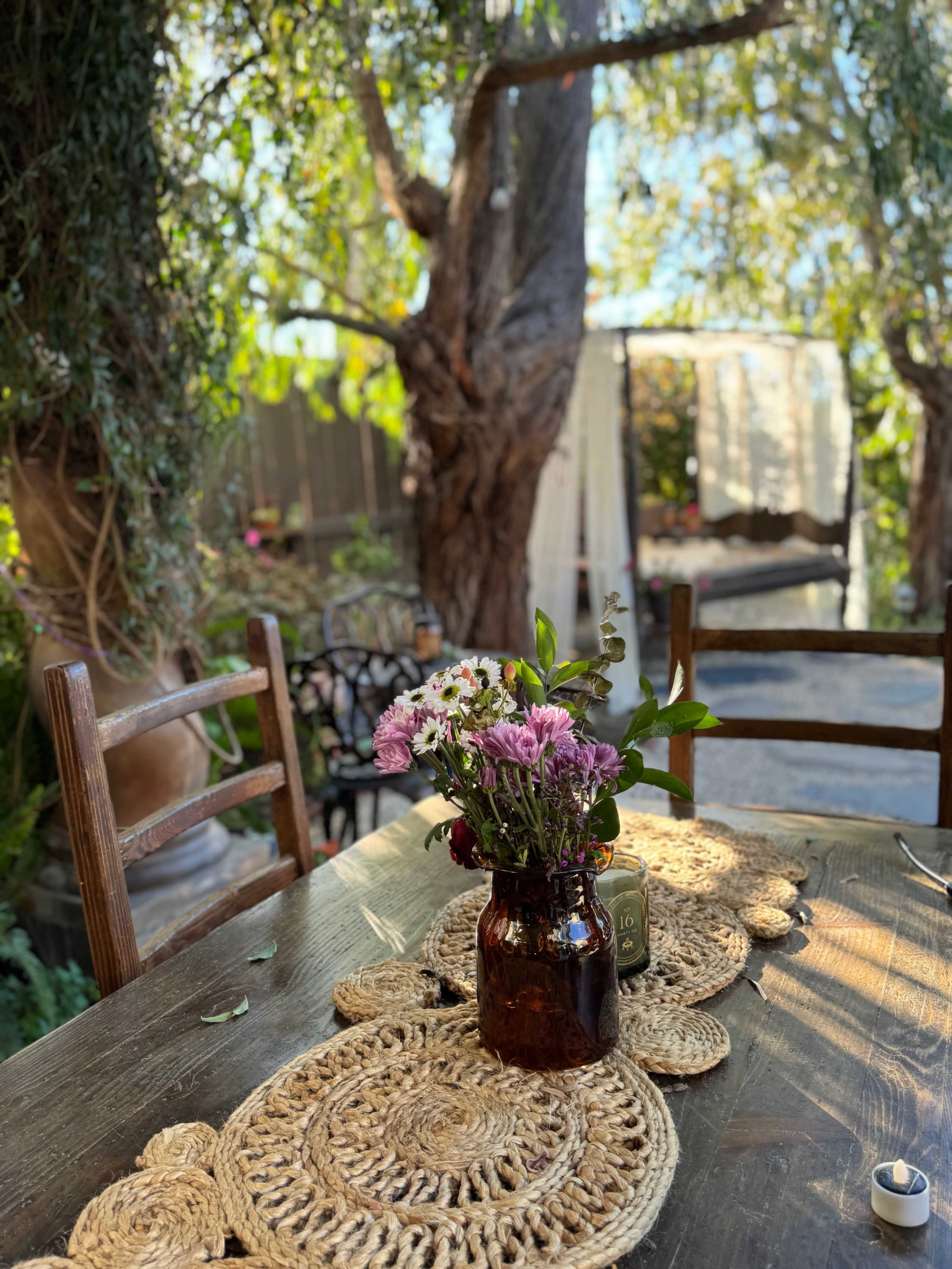 A wooden table is set outside, adorned with a woven placemat and a bouquet of wildflowers in a dark glass vase, surrounded by chairs and greenery.