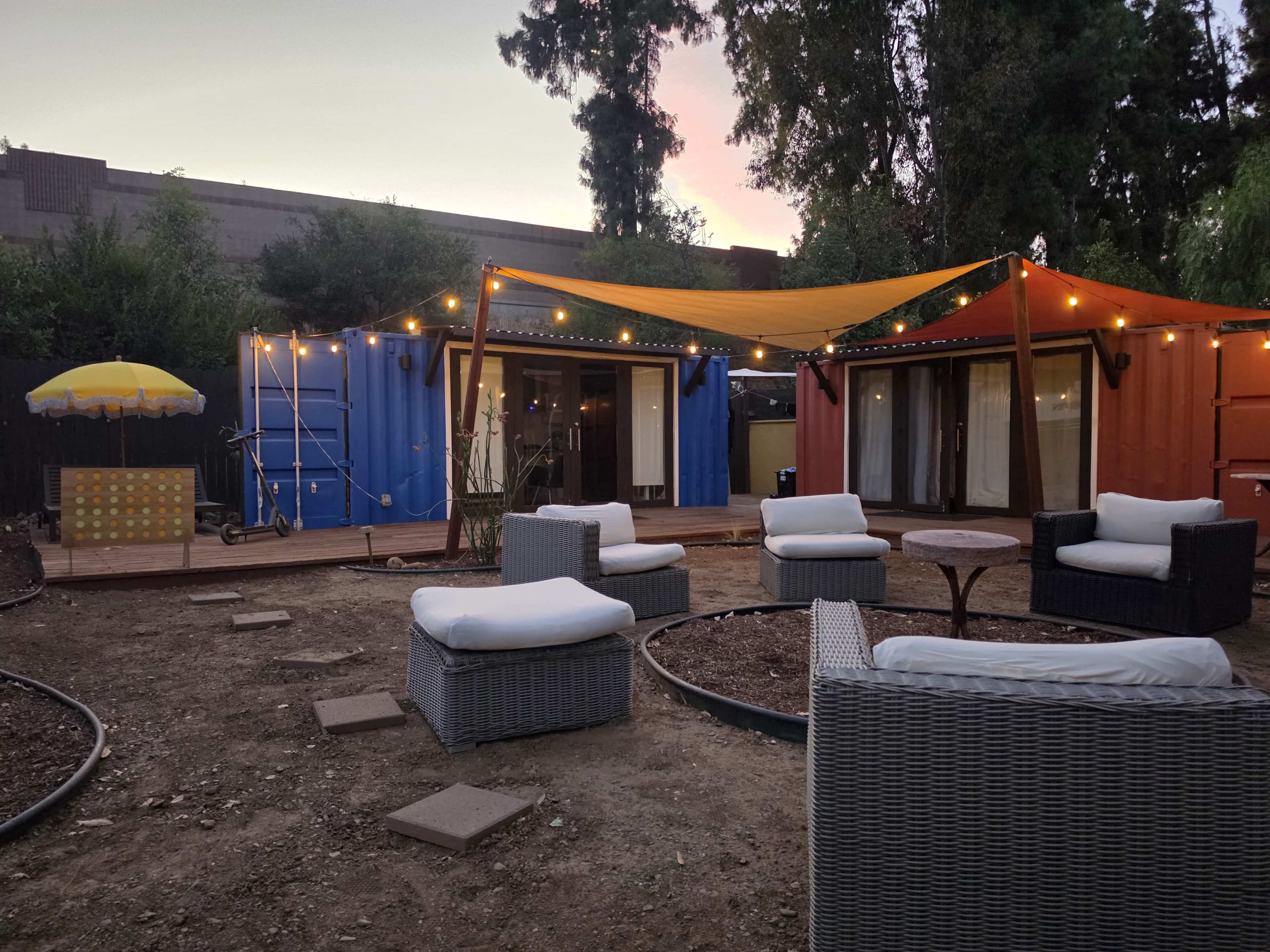 The image shows an outdoor seating area with several chairs and a table, surrounded by shipping container structures and decorative lights strung overhead.