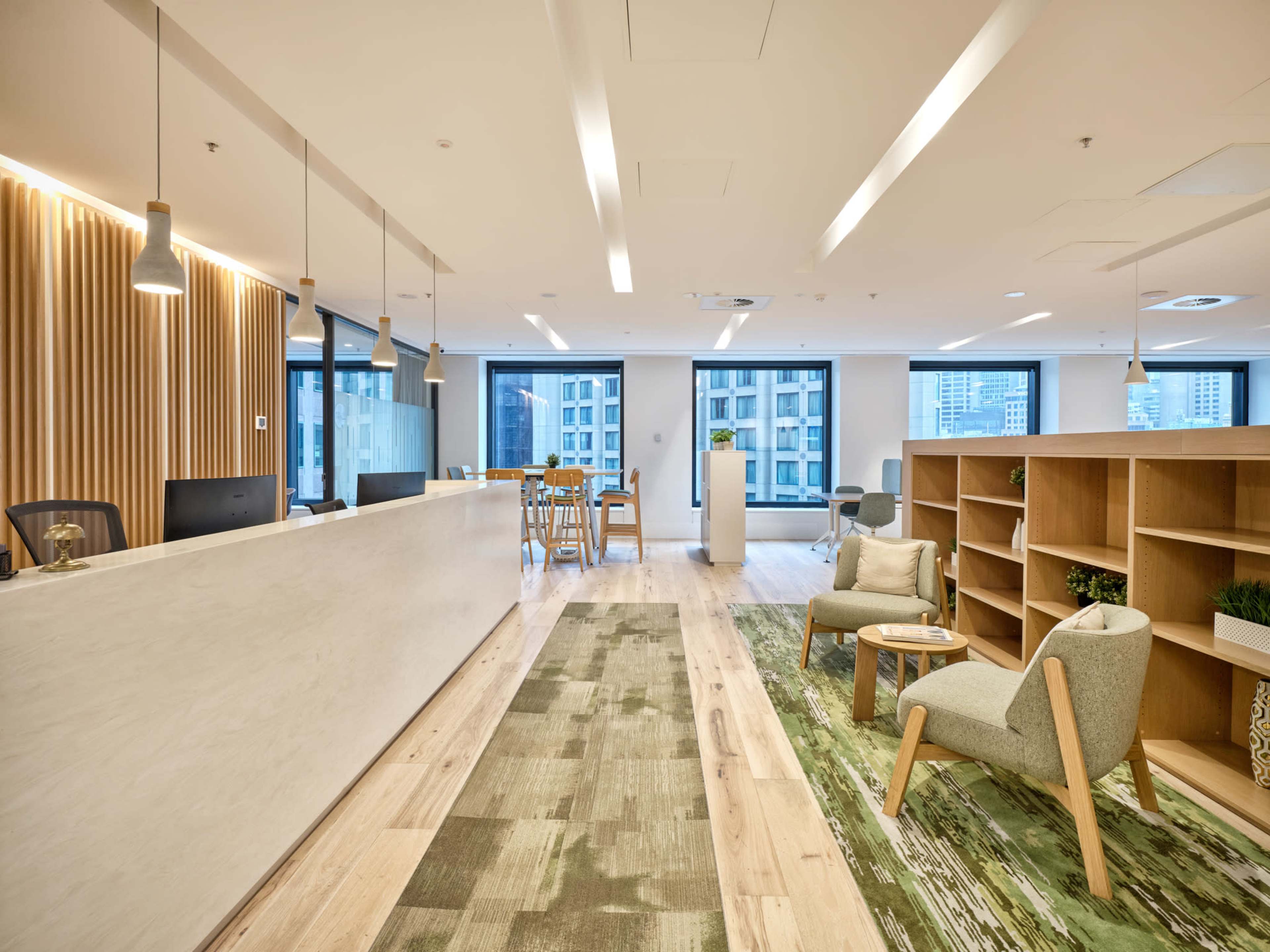 The image shows a modern office interior featuring a reception area with a white desk, wooden paneling, and green decor elements, along with seating and tables arranged near large windows.
