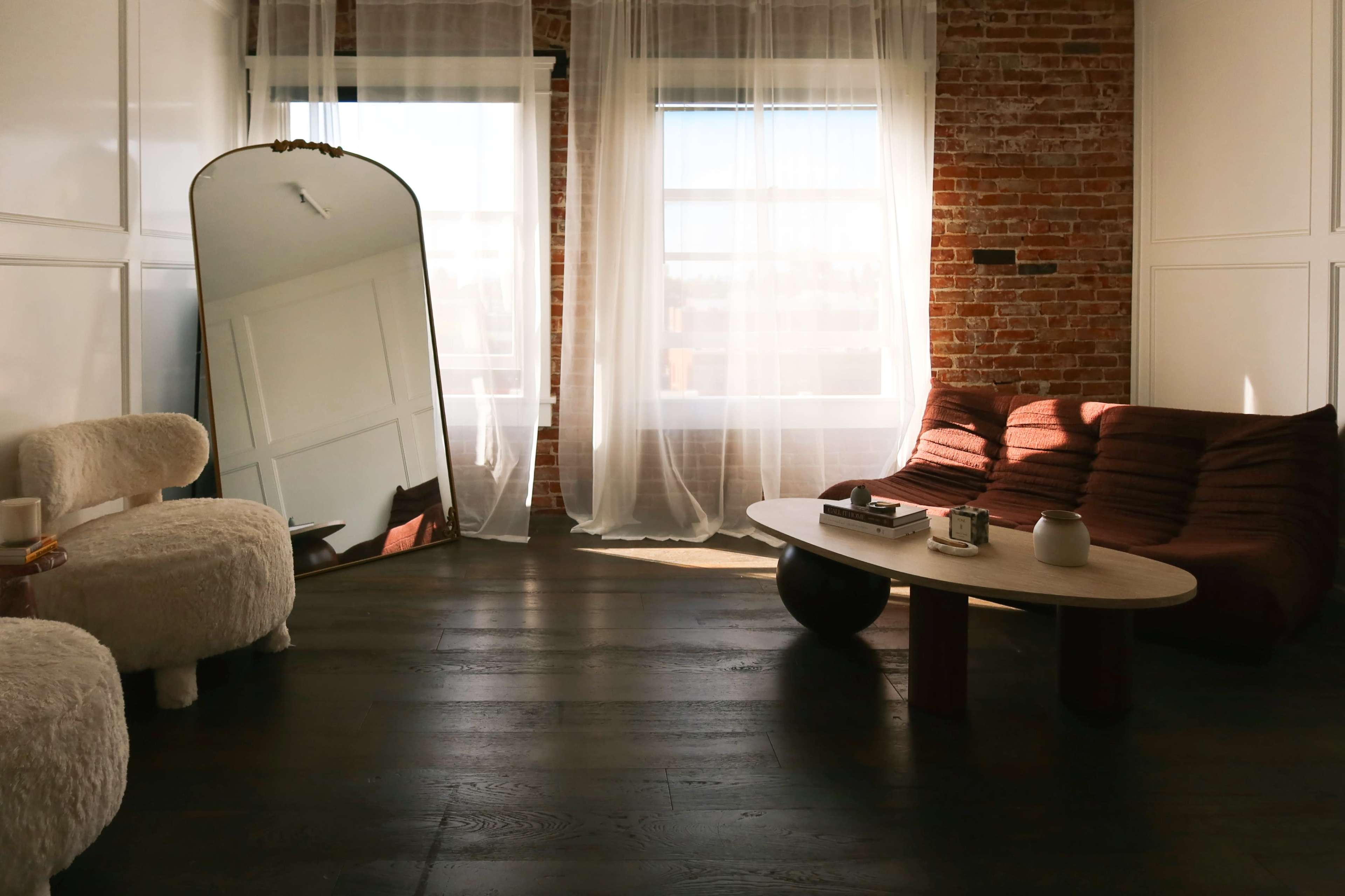 The room features exposed brick walls, a large mirror, a low coffee table, and a cozy seating area with natural light streaming through sheer curtains.