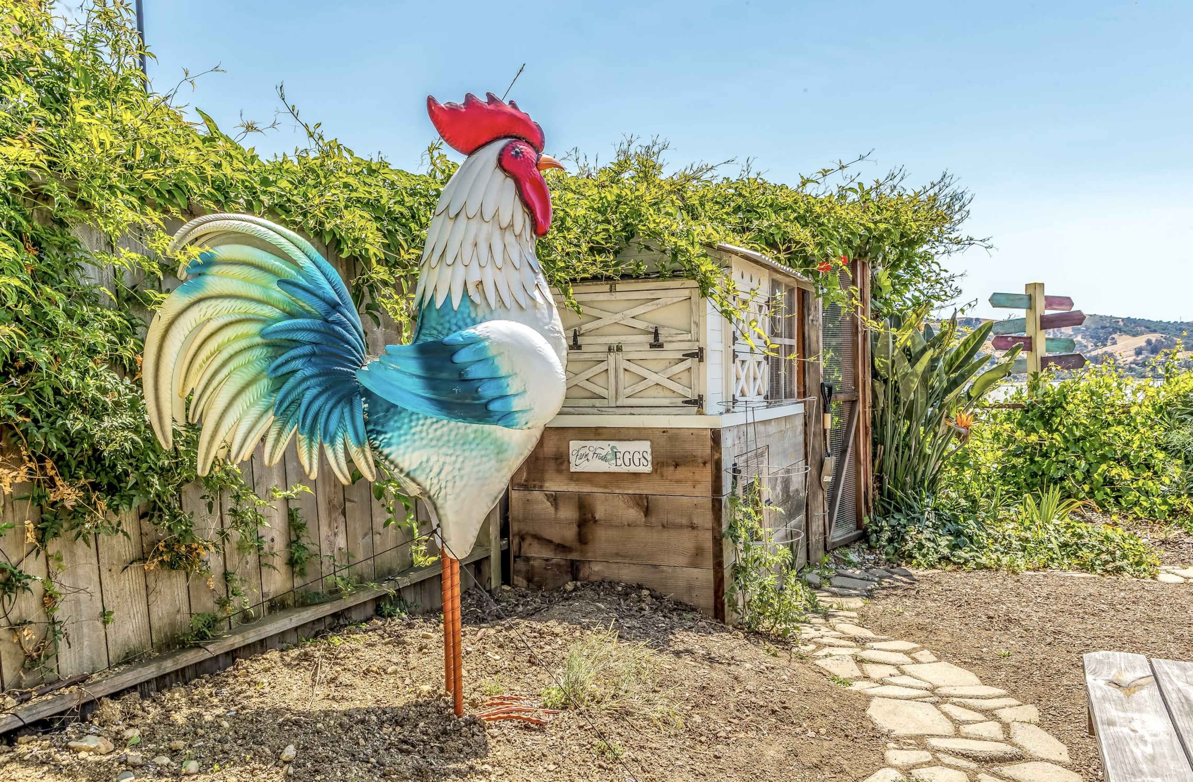 A large, colorful rooster sculpture stands in a garden near a wooden structure surrounded by greenery.