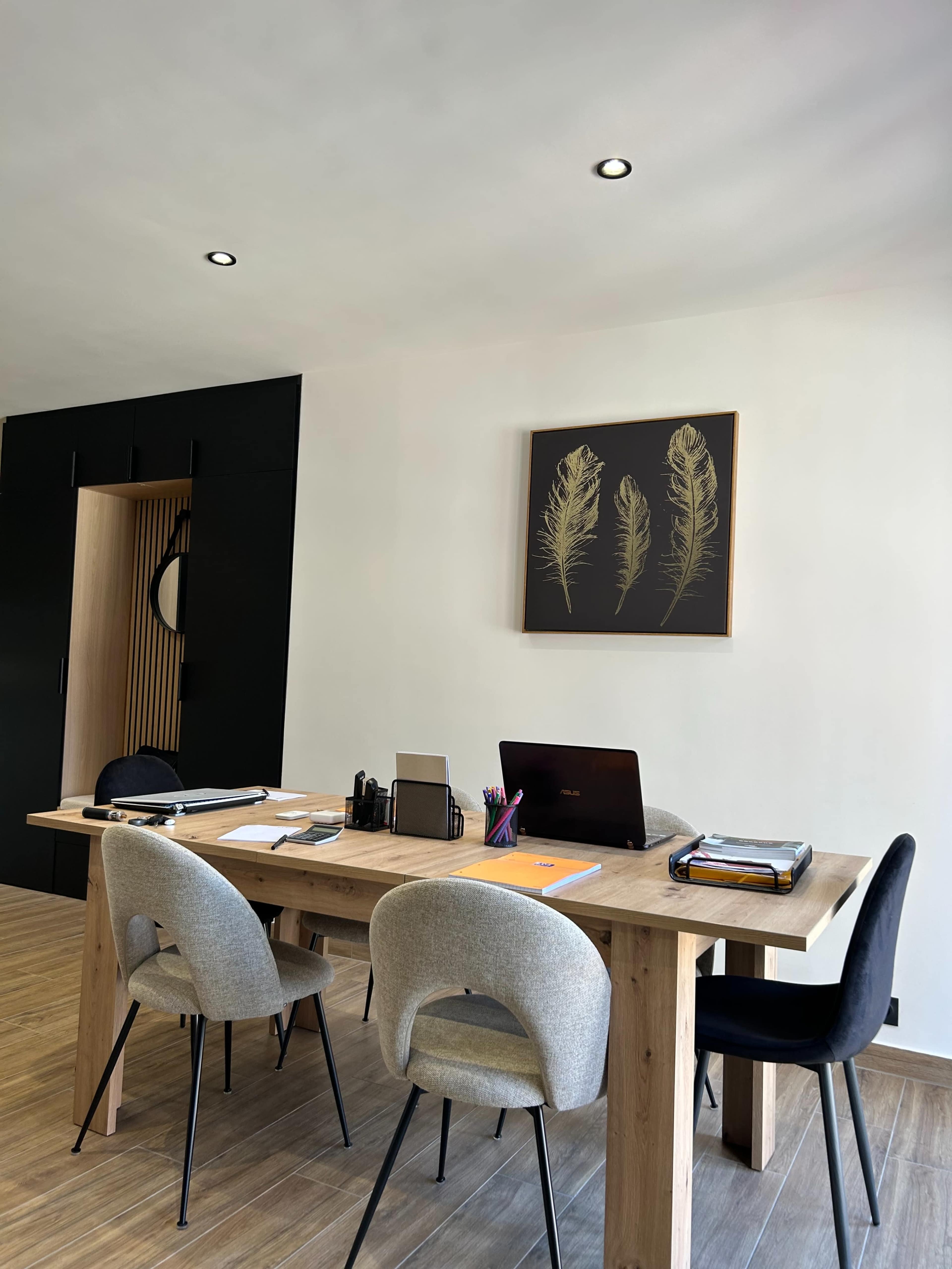 The image shows a modern office space with a wooden desk, three chairs, a laptop, and a framed artwork on the wall.
